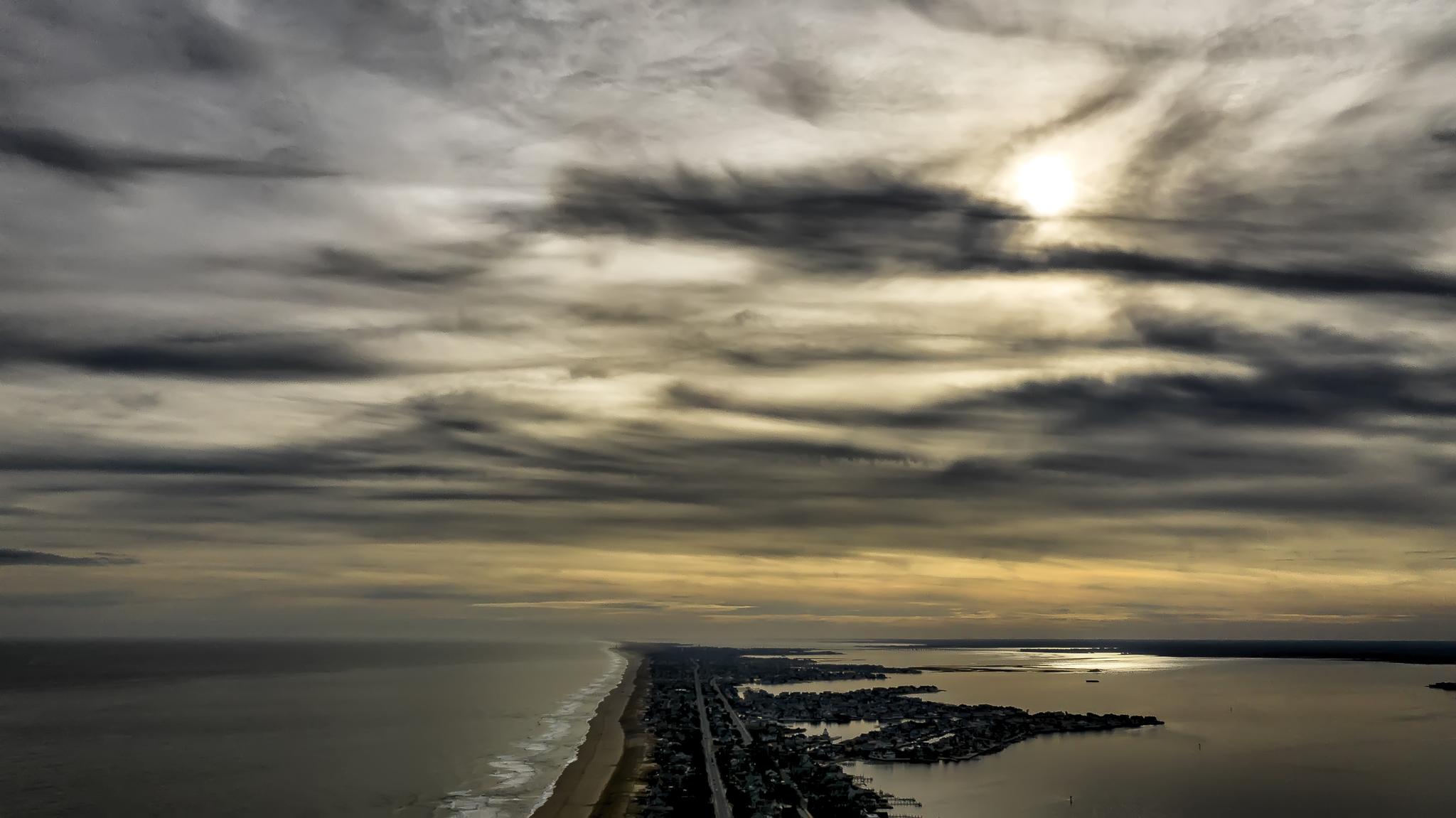Aerial view of a coastal strip between ocean and bay under dramatic cloudy sky.