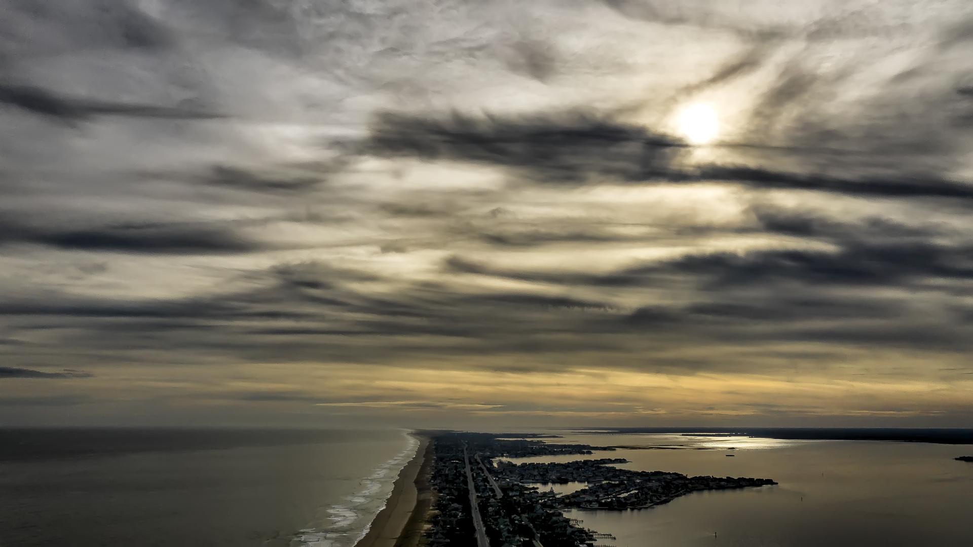 Aerial view of a coastal strip between ocean and bay under dramatic cloudy sky.