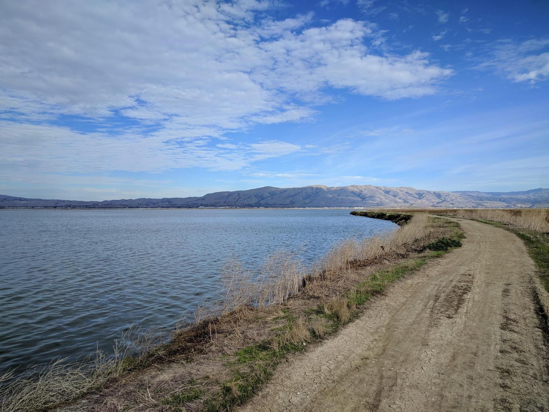 Sunny day with a dirt path beside calm blue water and distant mountains.