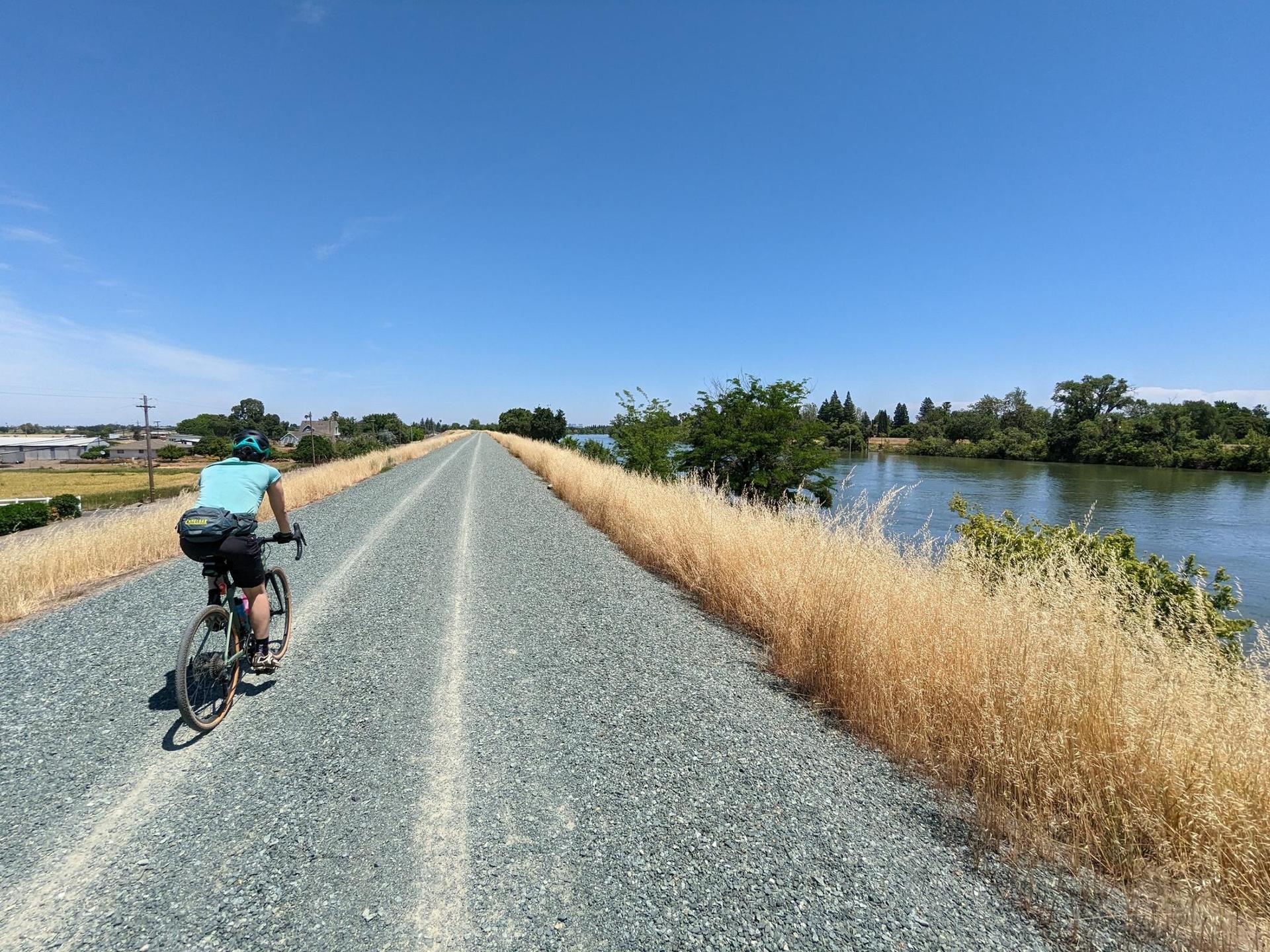 A person cycling on a gravel path next to a river under a bright blue sky.