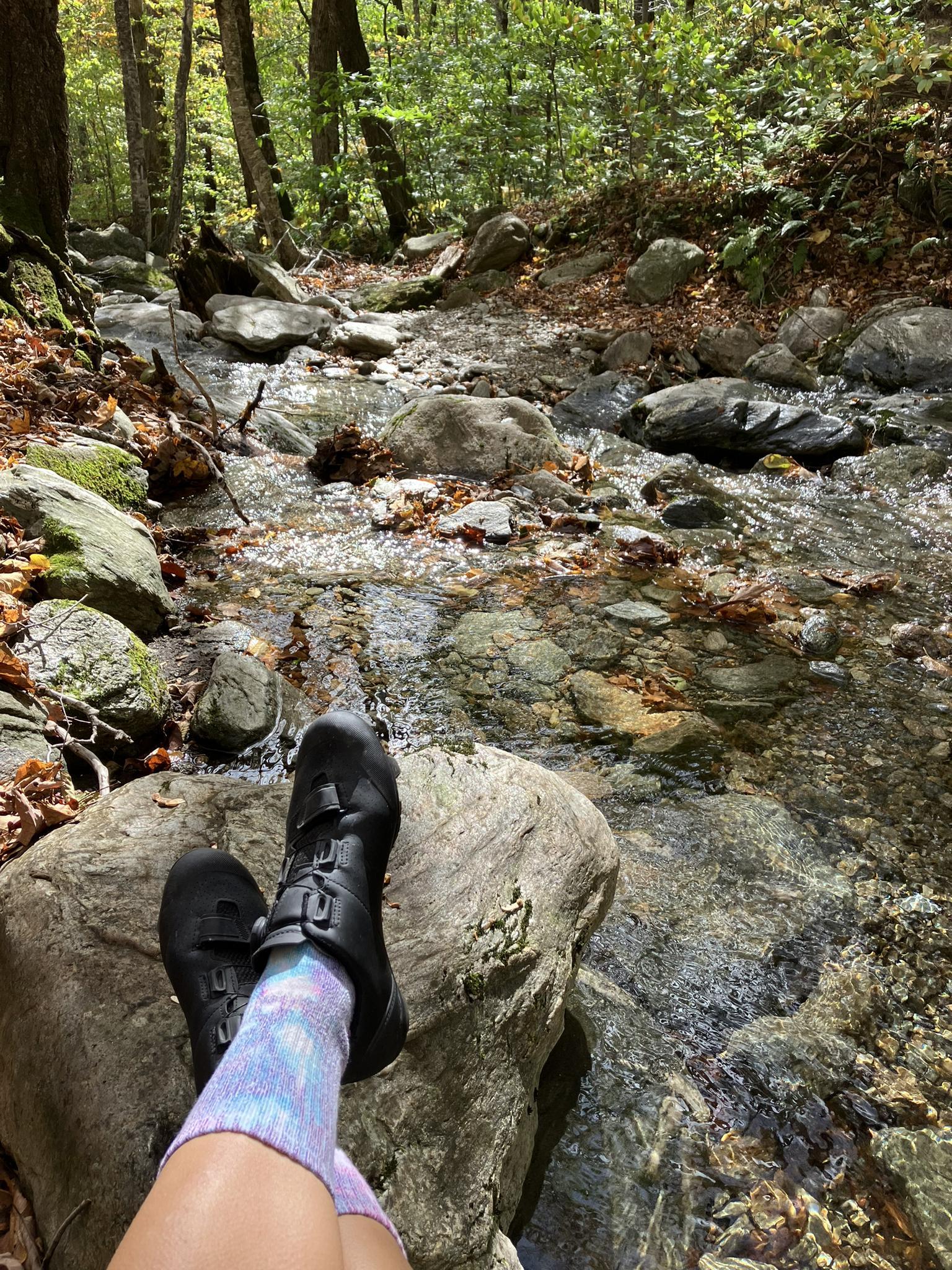 Feet in black boots and tie-dye socks resting by a sunlit forest stream.