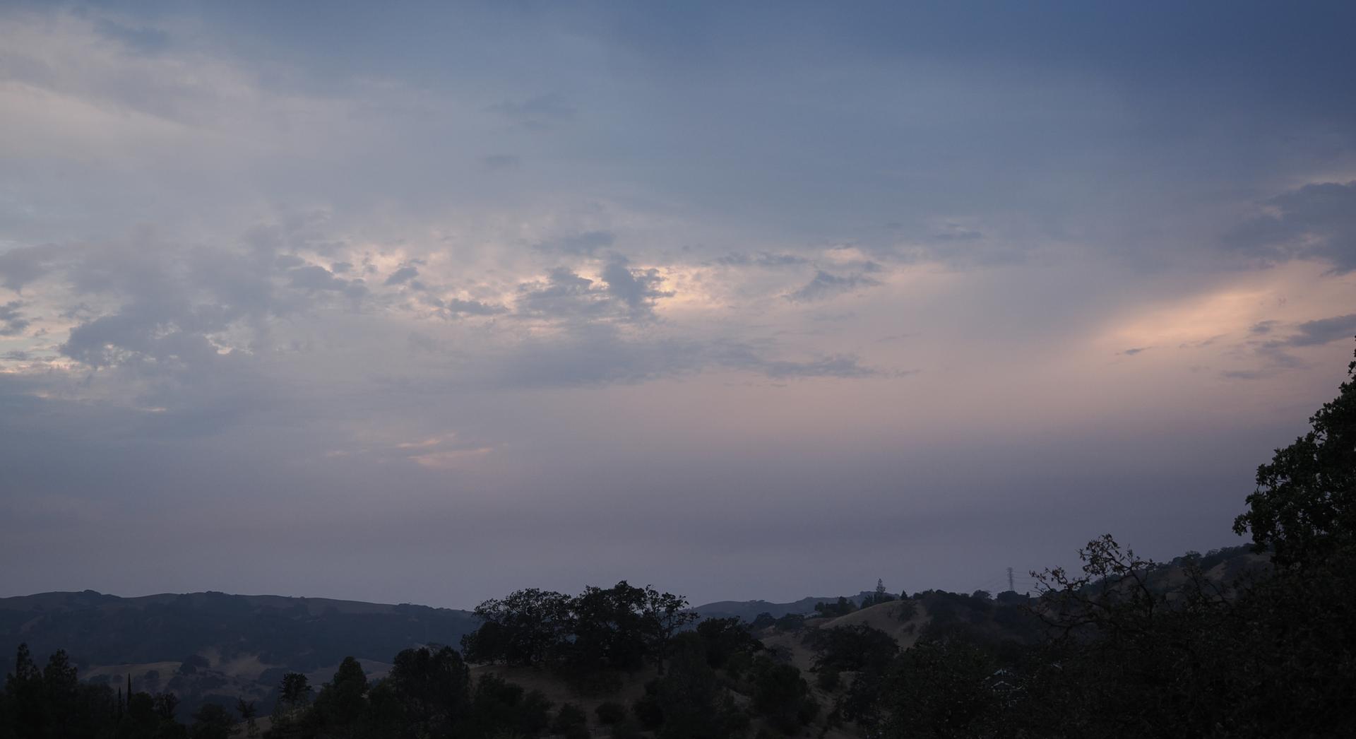 Cloudy sky with soft sunset colors over dark, rolling hills and trees.