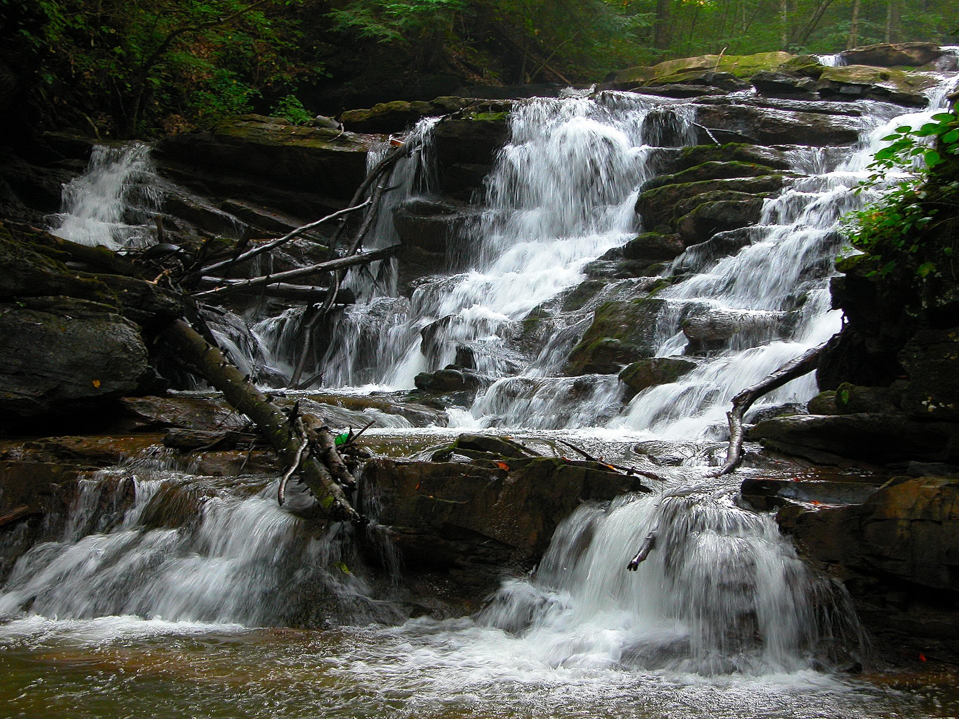 A multi-tiered waterfall cascades over dark rocks in a lush green forest.