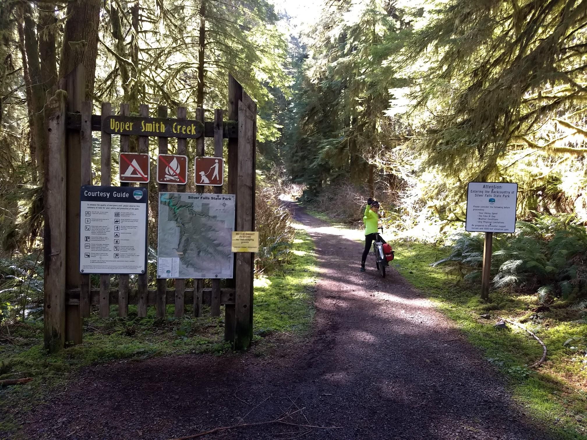 Person with bicycle on a sunny forest path next to an information board. - Newt Loop Trail – Smith Creek Village loop from 214 Trailhead
