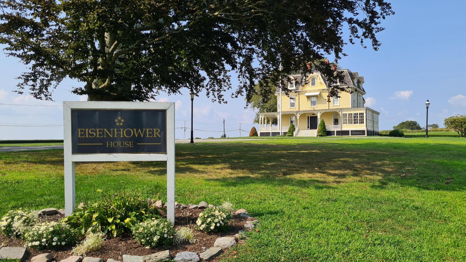 A sign stands in a green lawn with a large yellow house behind a tree.
