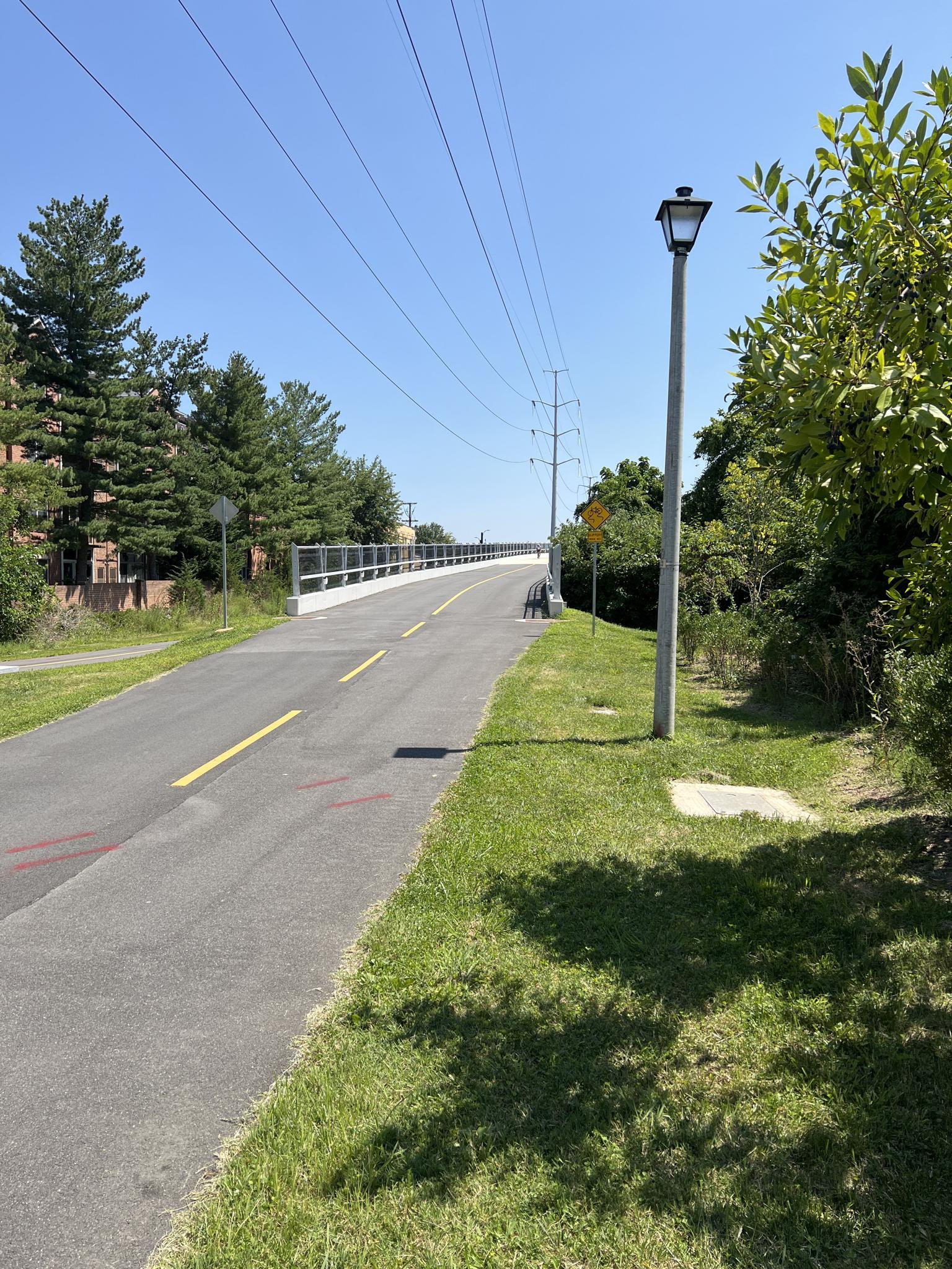 A sunny paved path with a bridge, lamppost, and power lines overhead.