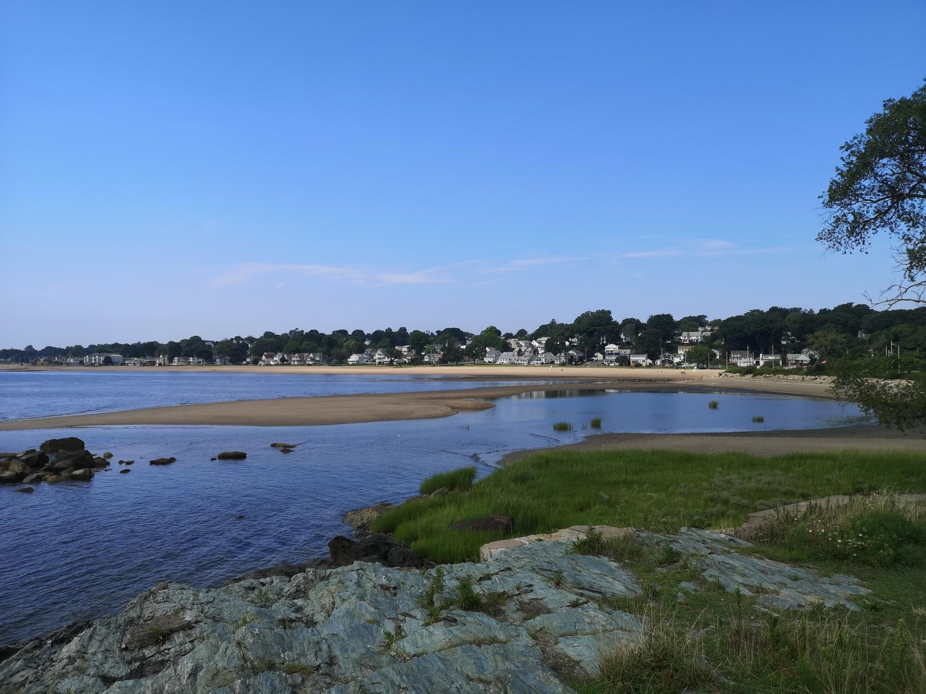 Rocky foreground overlooking a calm coastal inlet, sandy beach, and distant town.