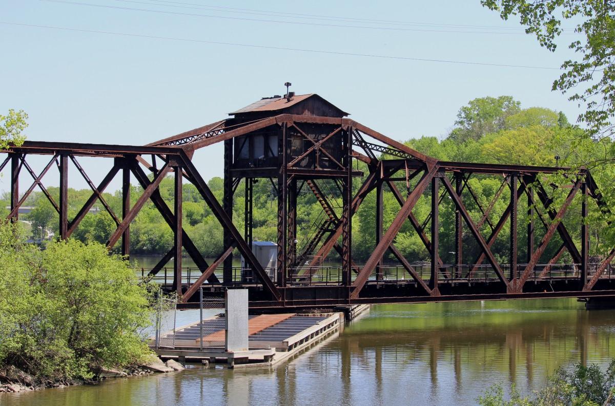 Rusty metal truss bridge spans a river with green trees, bright day.