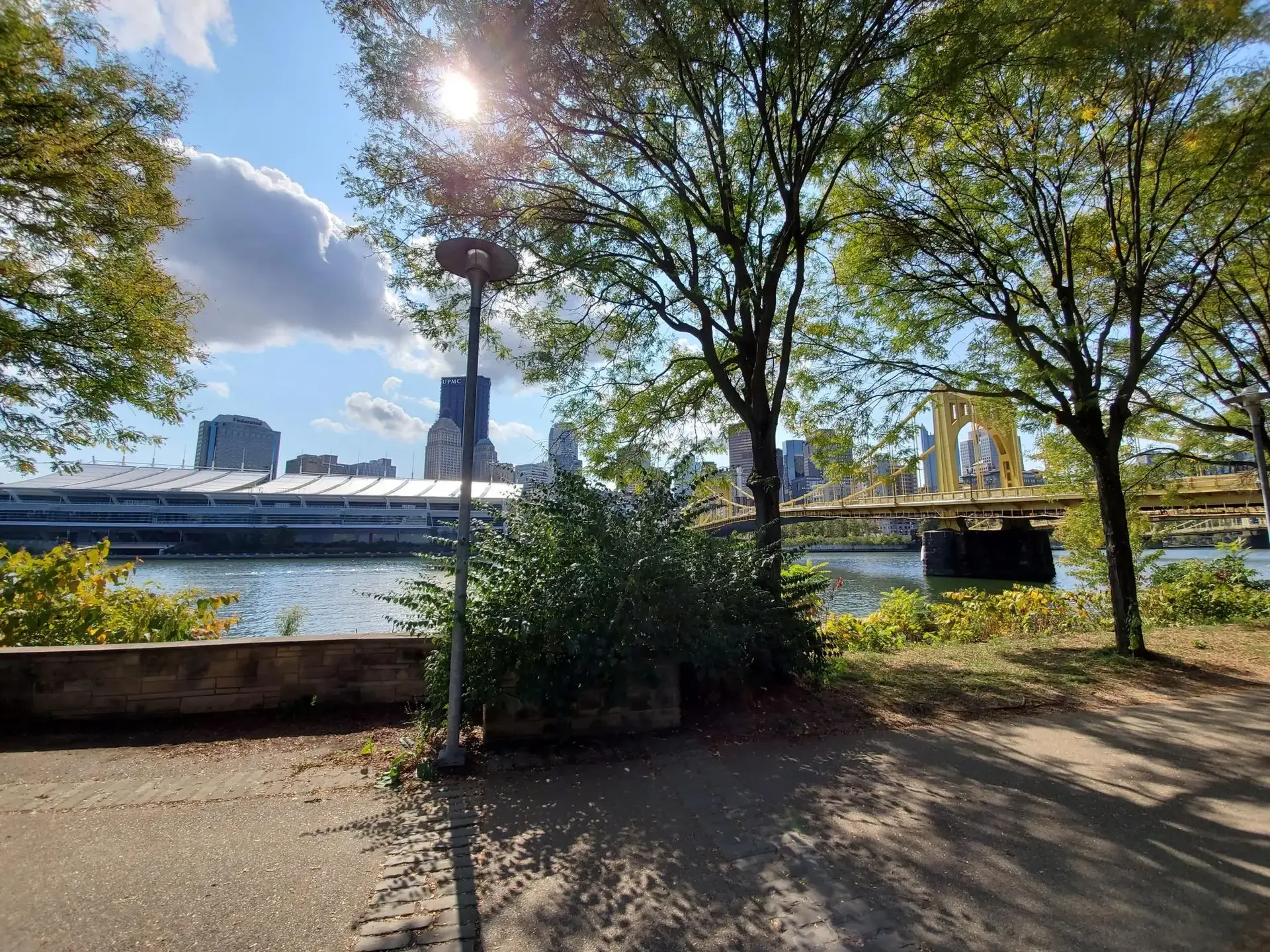 Sunny park path with trees, river, yellow bridge, and distant city buildings.