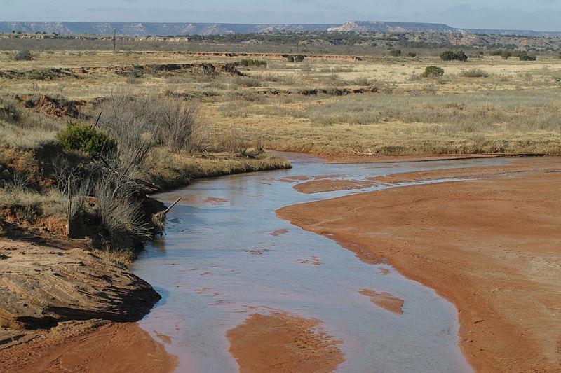 A winding muddy river flows through dry, grassy plains under a hazy sky.