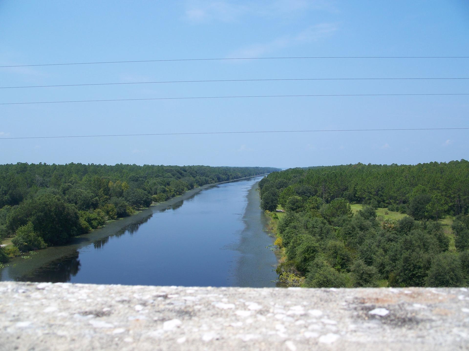 Straight river flowing through dense green forests under a clear blue sky.