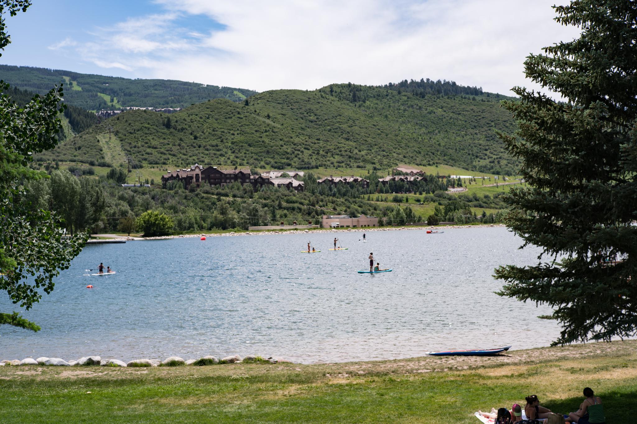People paddleboarding and relaxing by a lake with forested hills and buildings.