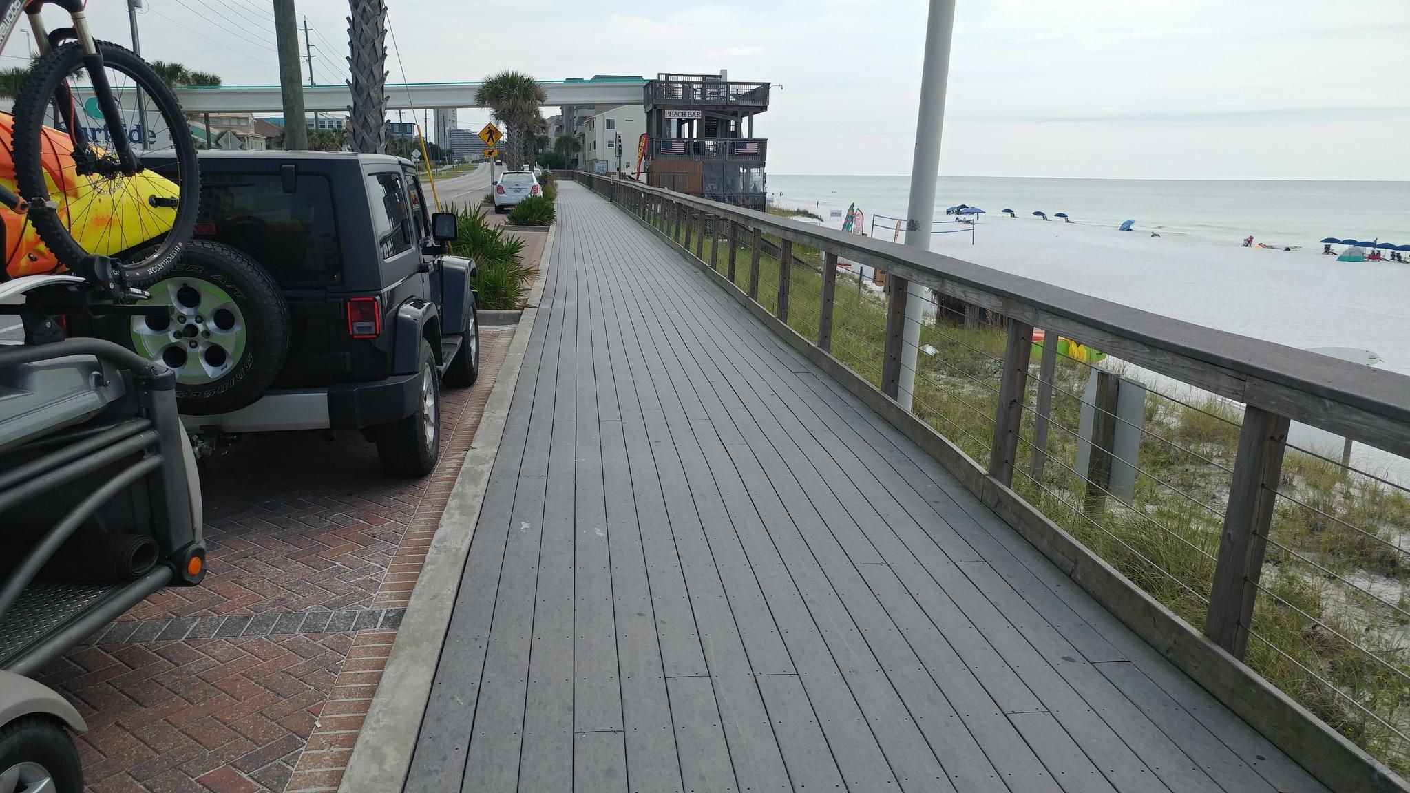 A boardwalk next to a sandy beach with a parked car and ocean under an overcast sky. - Scenic Highway 98 Trail – Destin Boardwalk loop from Destin