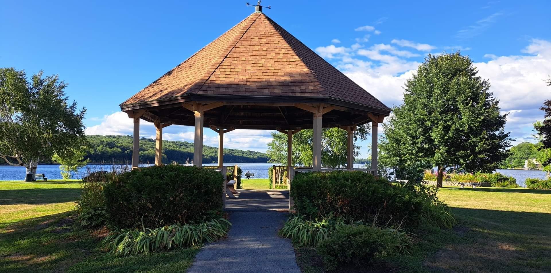 Wooden gazebo on a sunny day overlooking a lake with distant people and green grass.