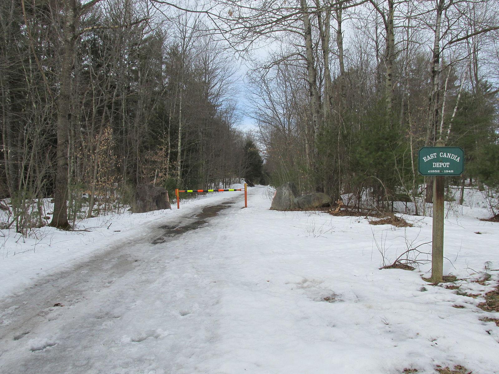 A snow-covered dirt road with a barrier and sign leading into a winter forest.