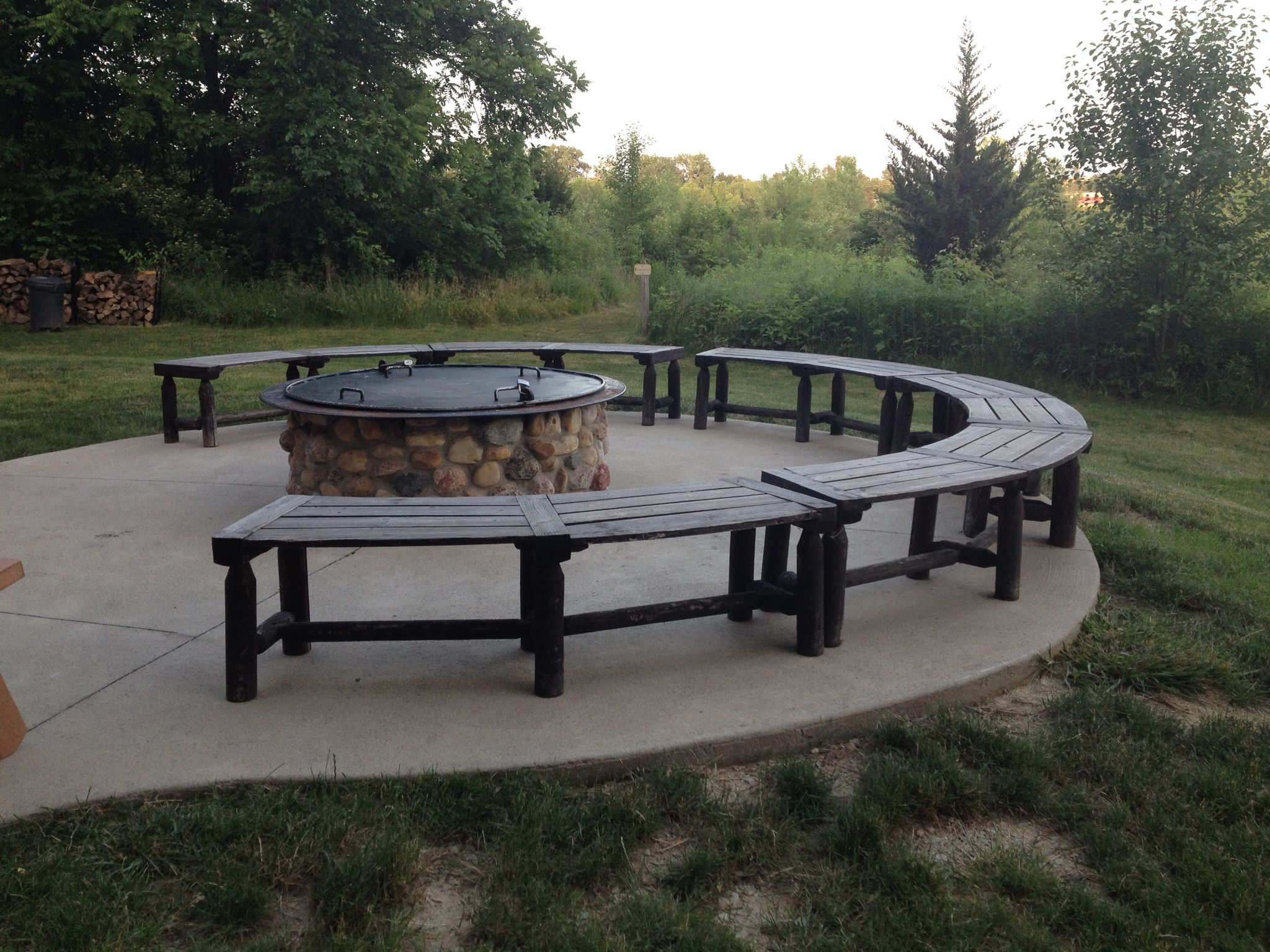 Outdoor circular stone fire pit and curved wooden benches on a patio, surrounded by trees. - Ritchey Woods Nature Preserve loop run