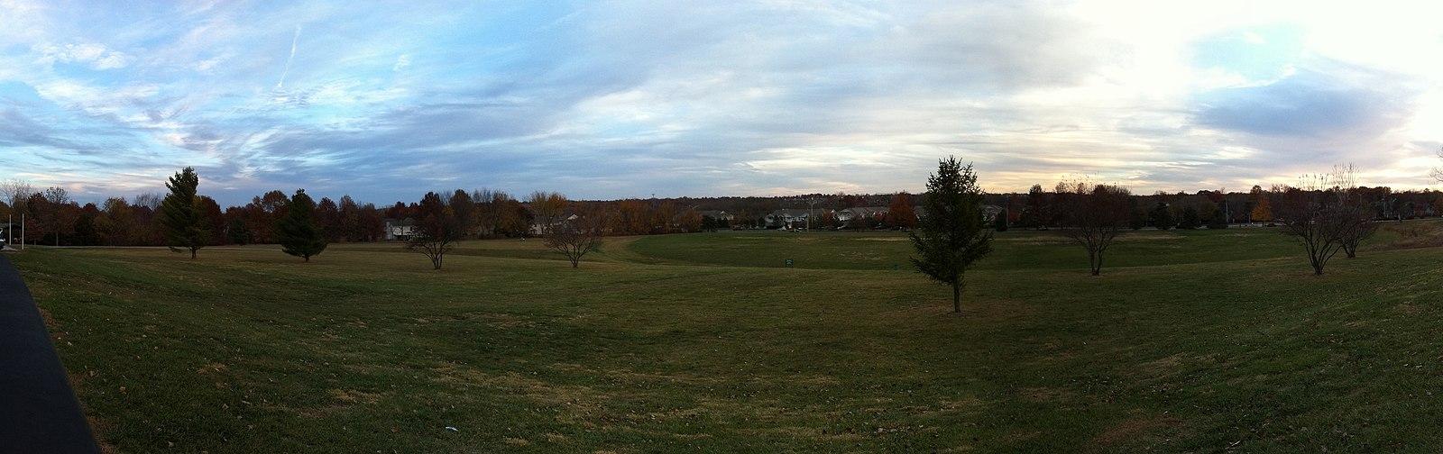 Panoramic view of a wide green field with scattered trees under a cloudy sky.
