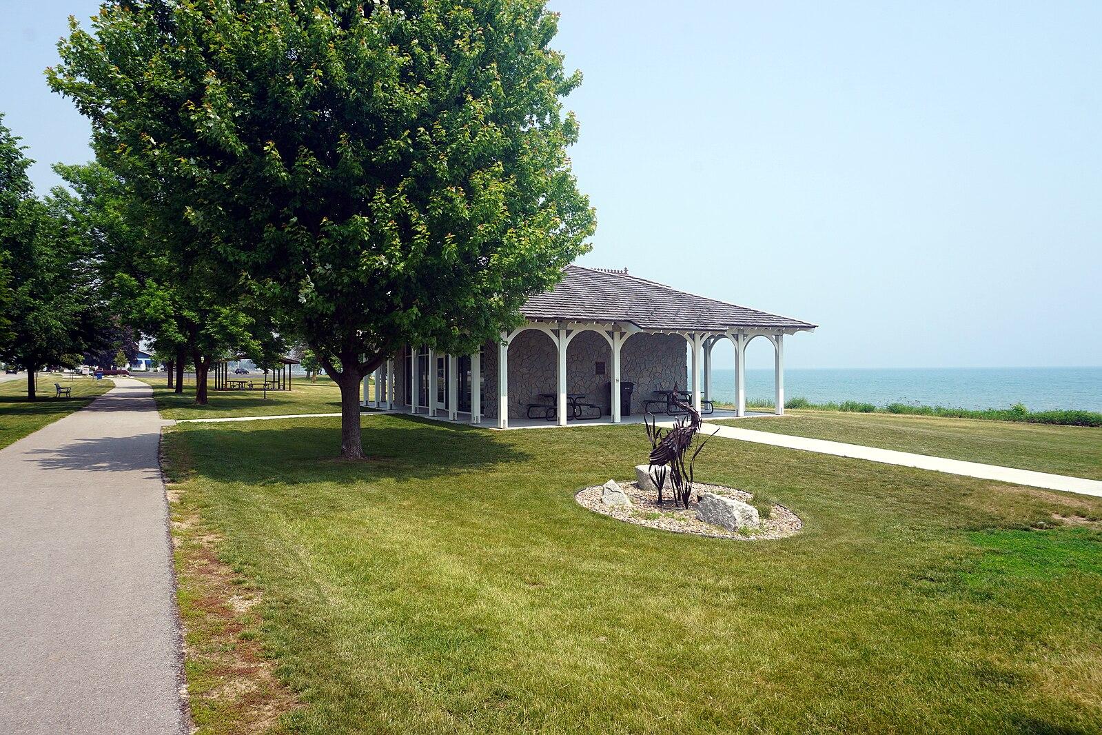 Sunny park with a white pavilion, green trees, and a distant body of water.