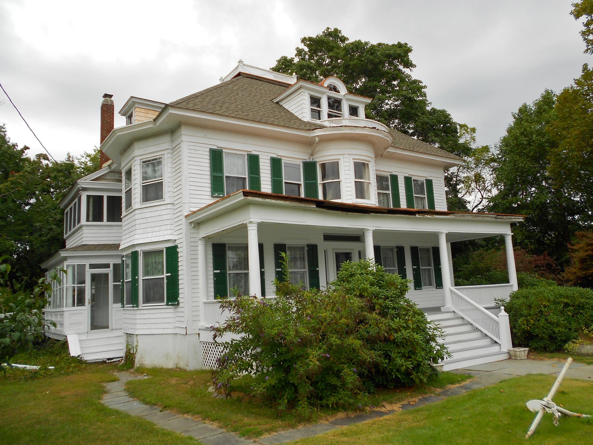 A large white house with green shutters and a porch, surrounded by greenery.