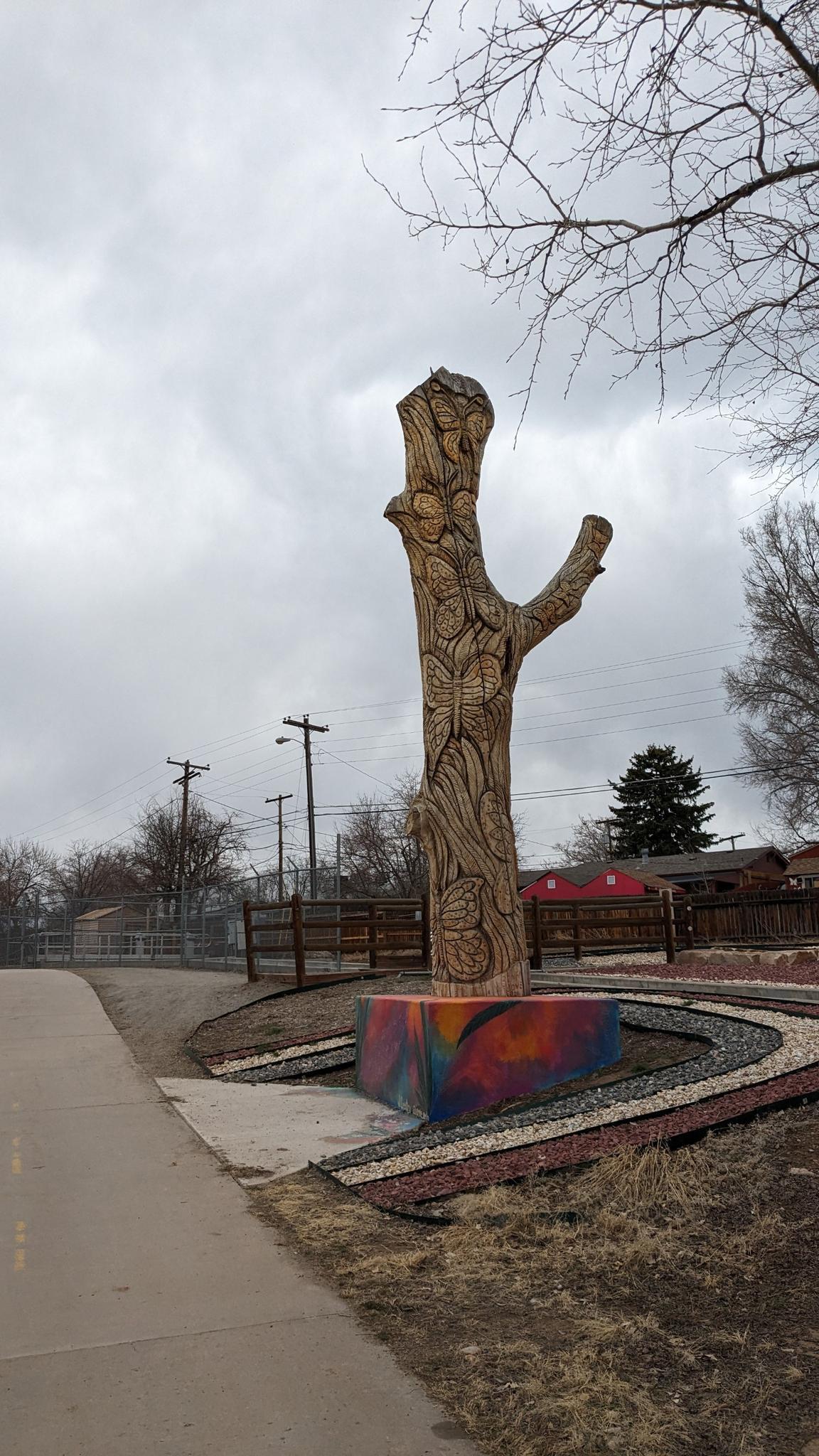 Tall carved wooden sculpture next to a paved path on a cloudy day.