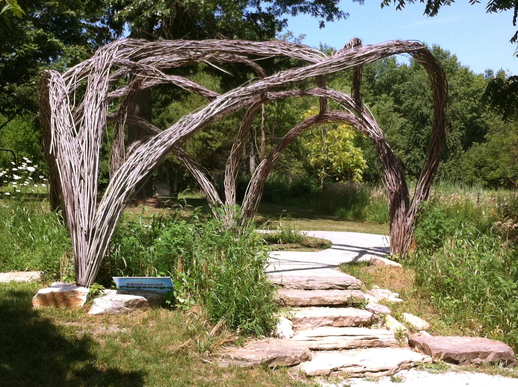 Woven branch archway over stone steps in a sunny, green outdoor setting.