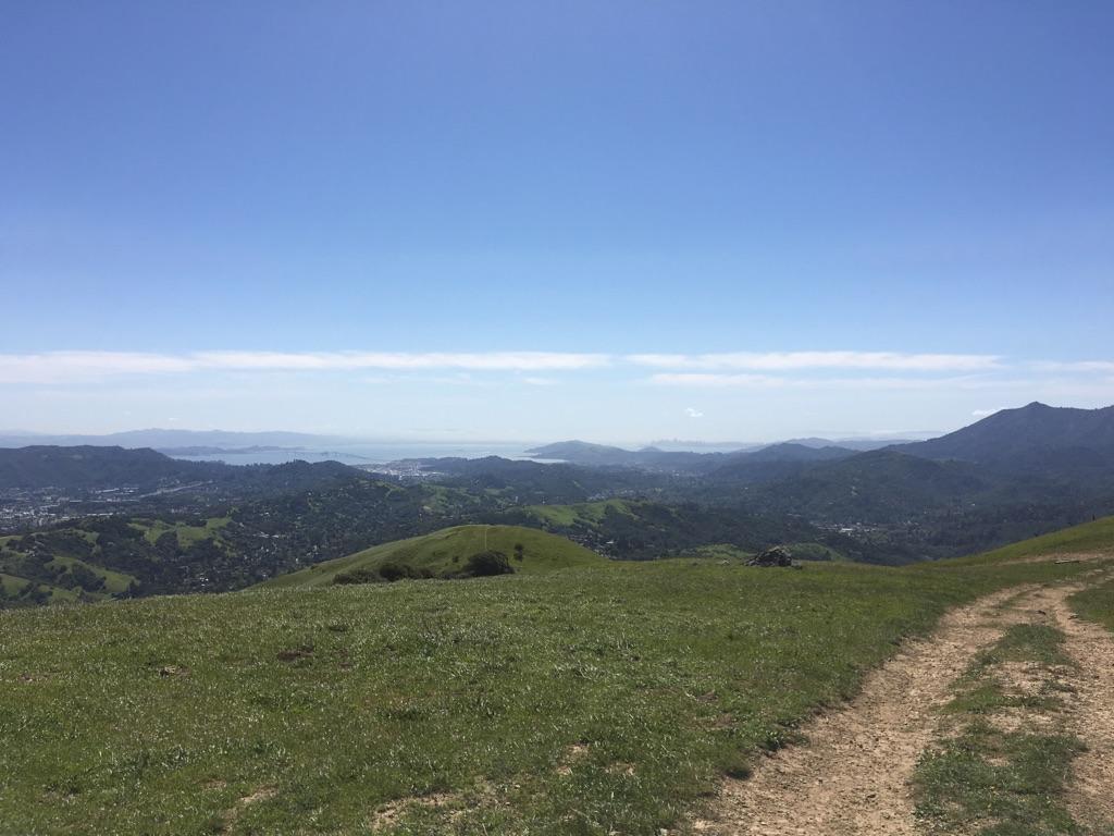 Expansive view of rolling green hills, distant mountains, and a dirt path under a clear sky.