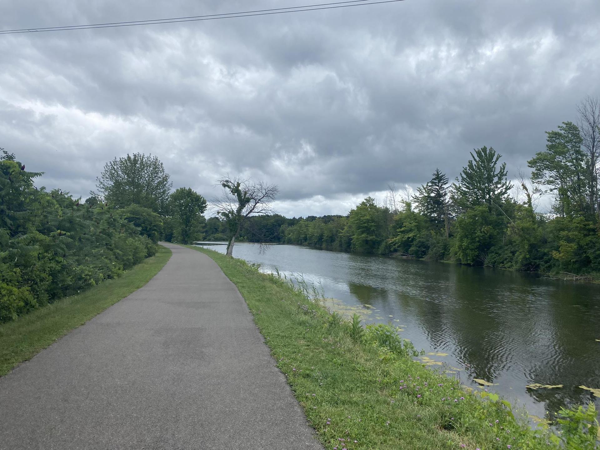 Paved path alongside a wide canal with green trees under a cloudy sky.