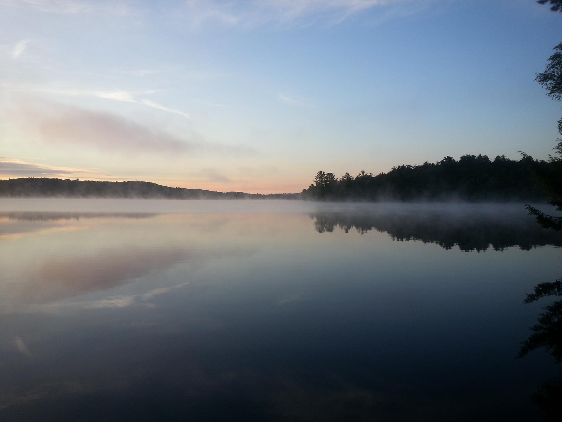 Serene misty lake at dawn with forested shores under a clear sky.