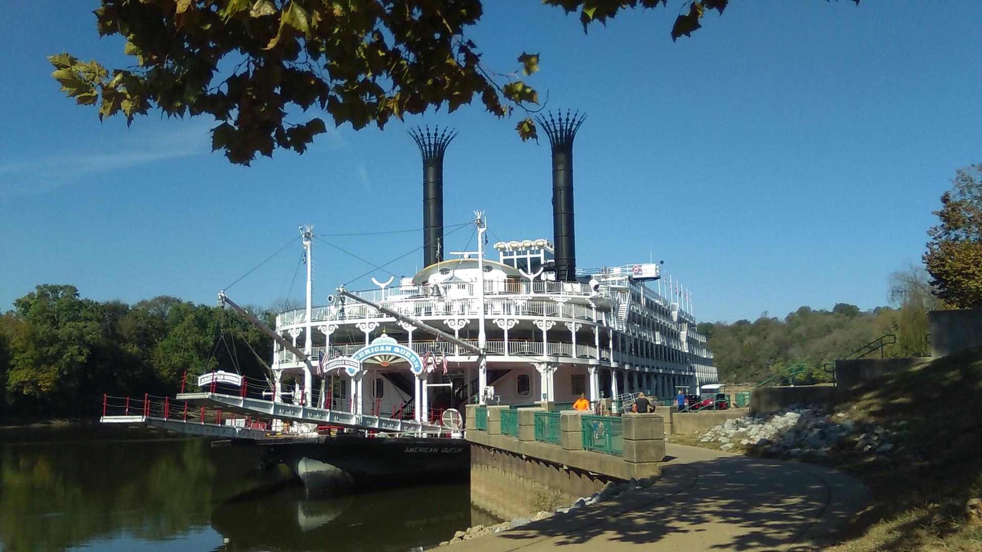 Large white paddlewheel boat docked on a sunny river with green trees.
