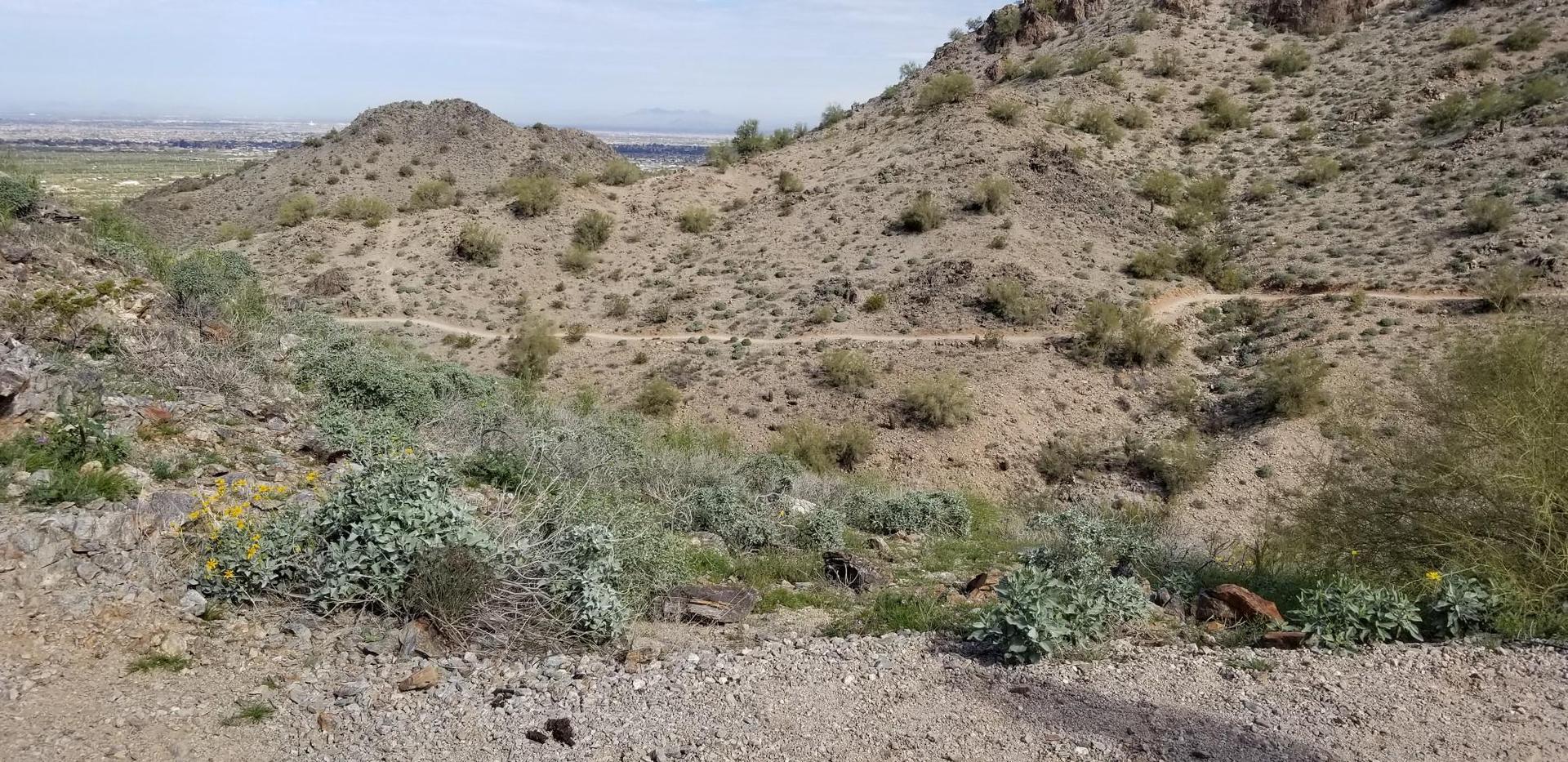 A winding dirt path traverses arid, rocky hills under bright daylight.