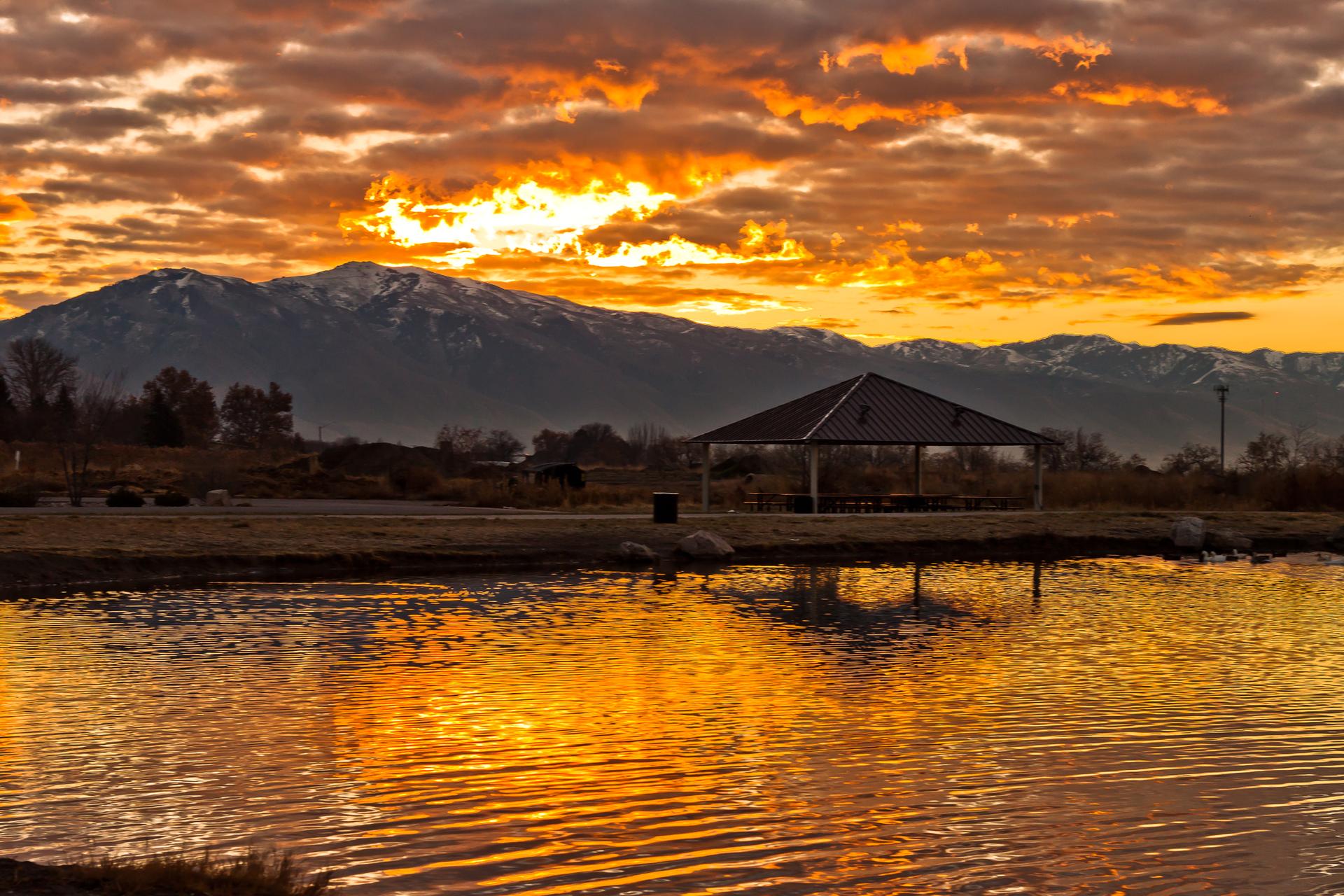 A covered pavilion by a lake reflecting a dramatic orange sunset over mountains.