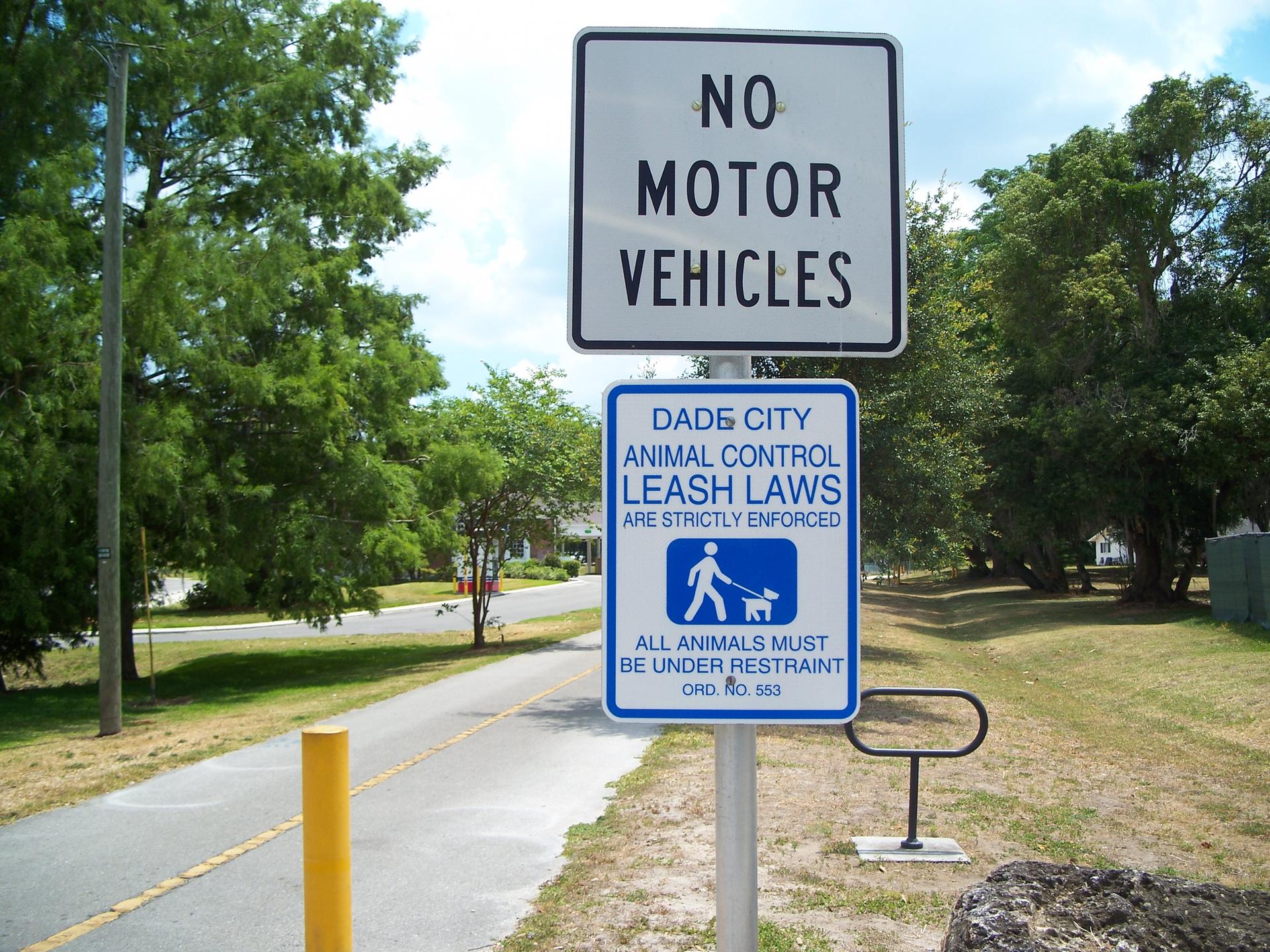 Two regulatory signs on a pole beside a paved path, surrounded by green trees.