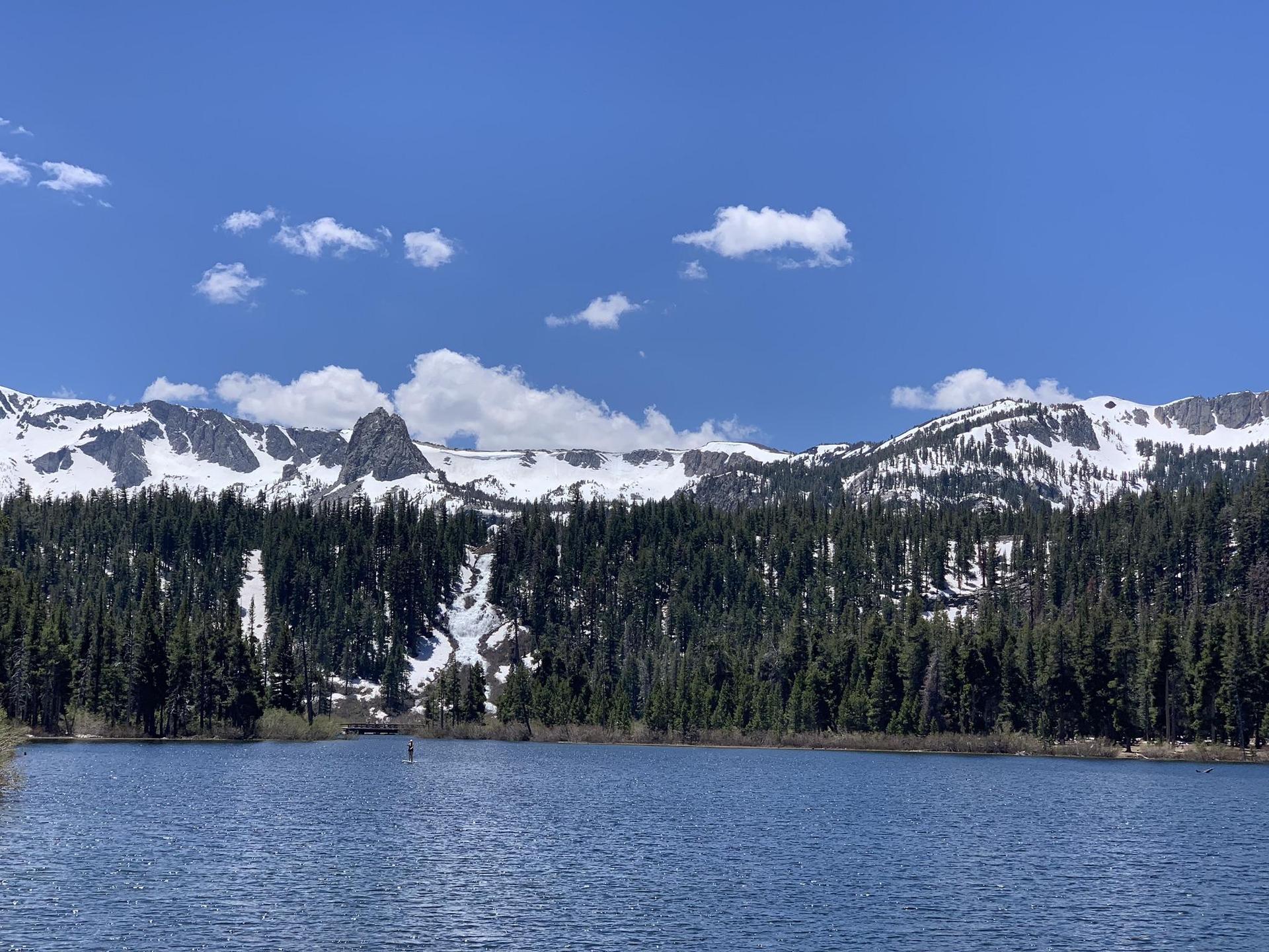 Calm blue lake reflecting snow-capped mountains and a pine forest.