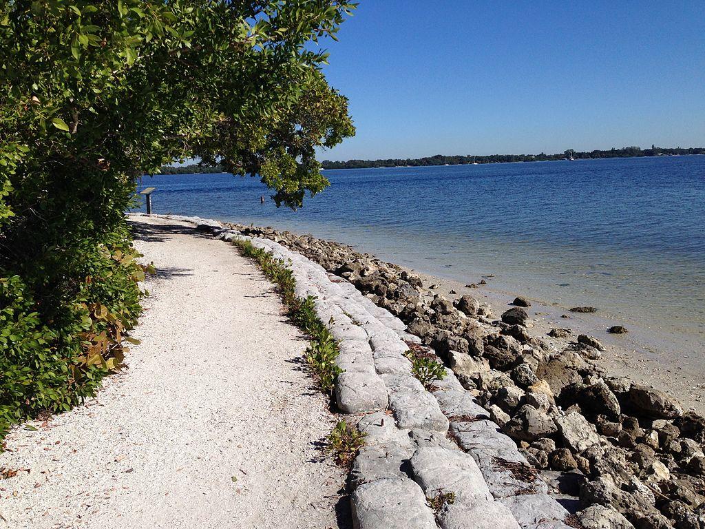 Sunny gravel path curves along a rocky shore next to calm blue water.