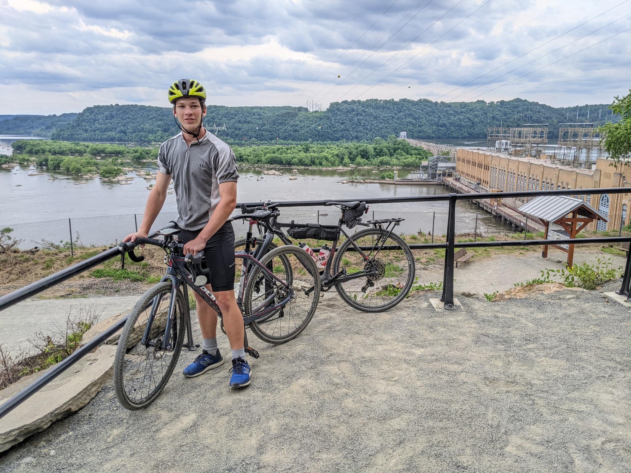 Young man with two bikes overlooking a river and dam under cloudy skies.