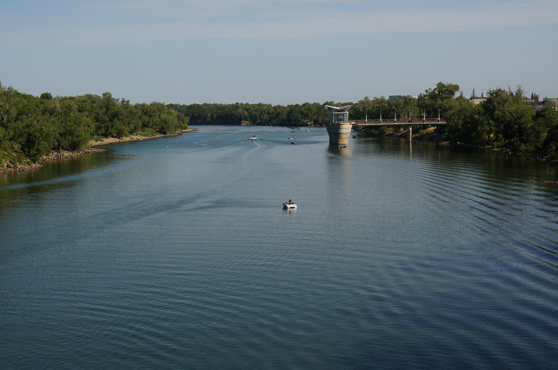 A small boat on a wide river with forested banks and a distant bridge.