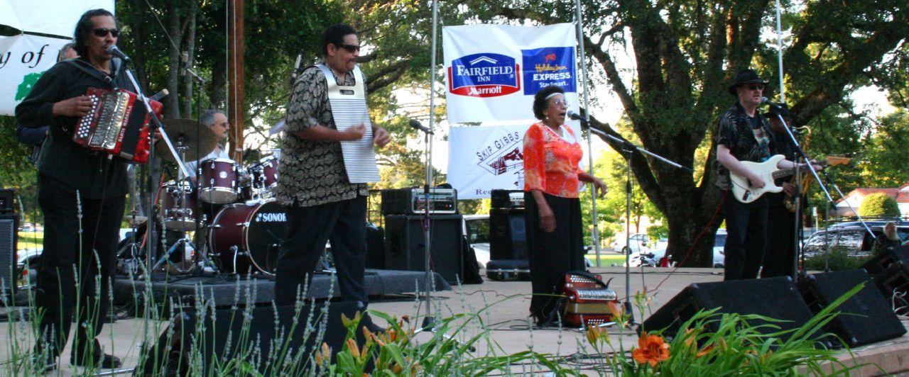 Four musicians perform on an outdoor stage with instruments and microphones.