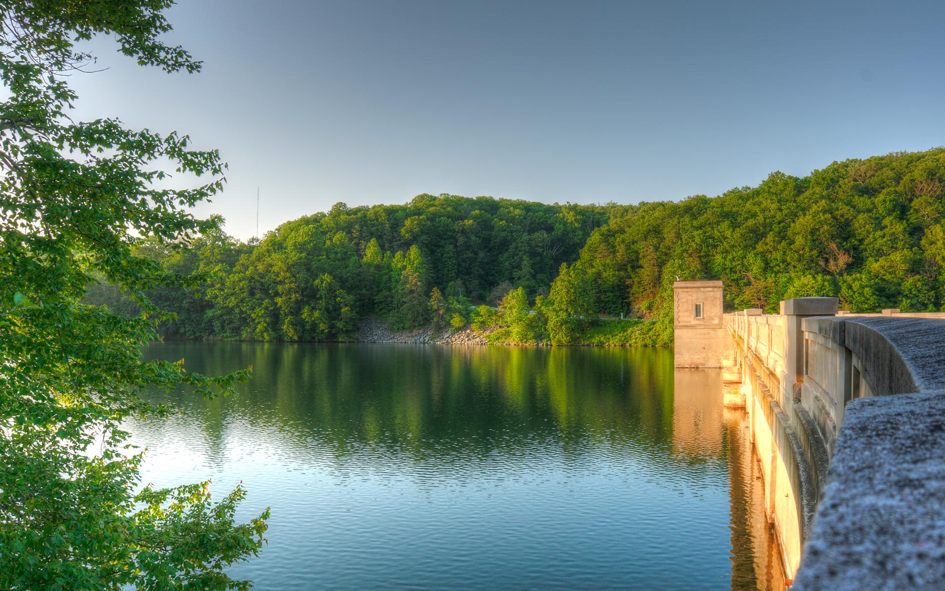 A serene lake next to a concrete dam, surrounded by lush green forests under clear skies.