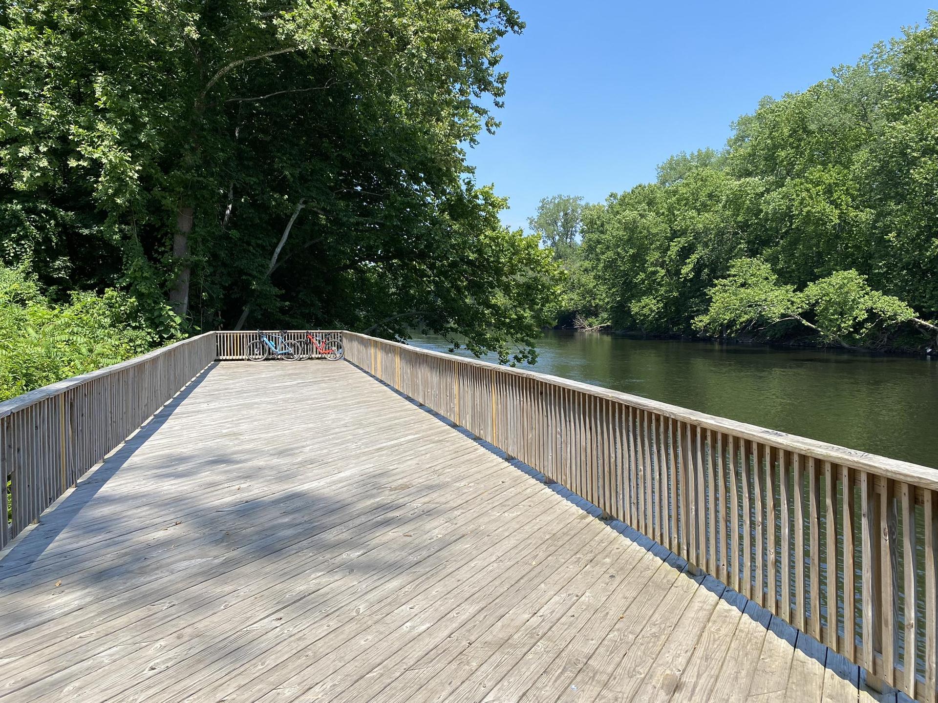 A long wooden boardwalk extends beside a calm river with lush green trees.