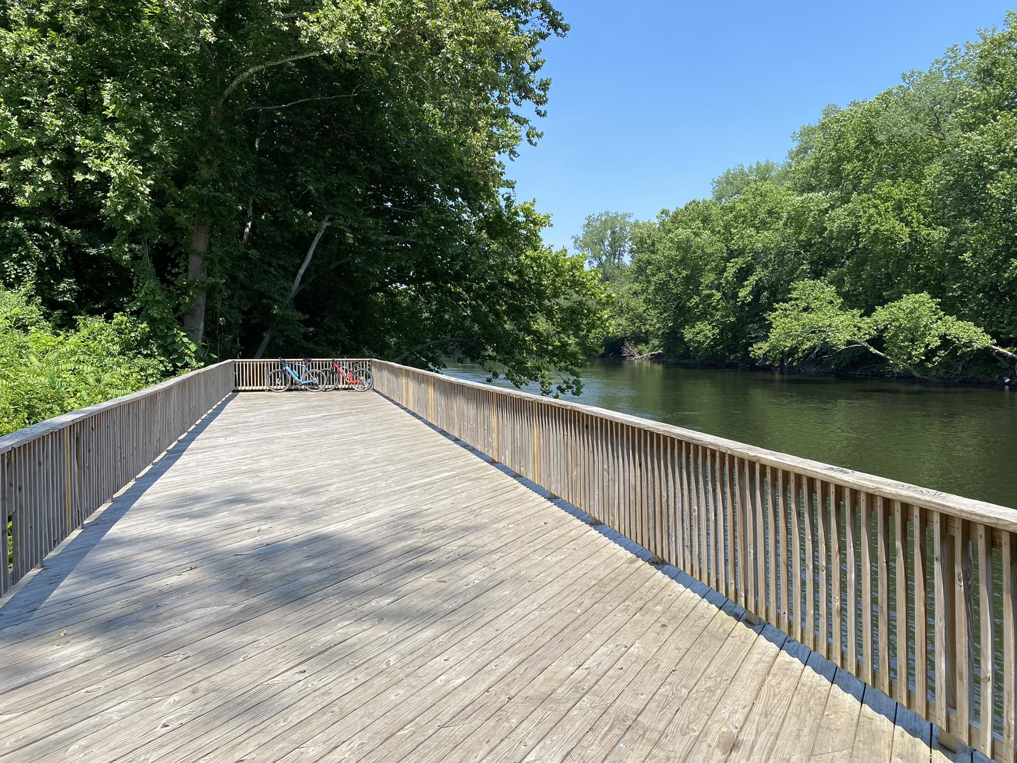 A long wooden boardwalk extends beside a calm river with lush green trees. - Two Rivers Trailway Loop