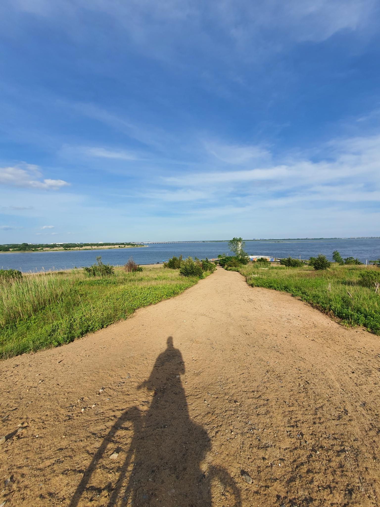 Cyclist's shadow on a sunny dirt path overlooking a wide bay.