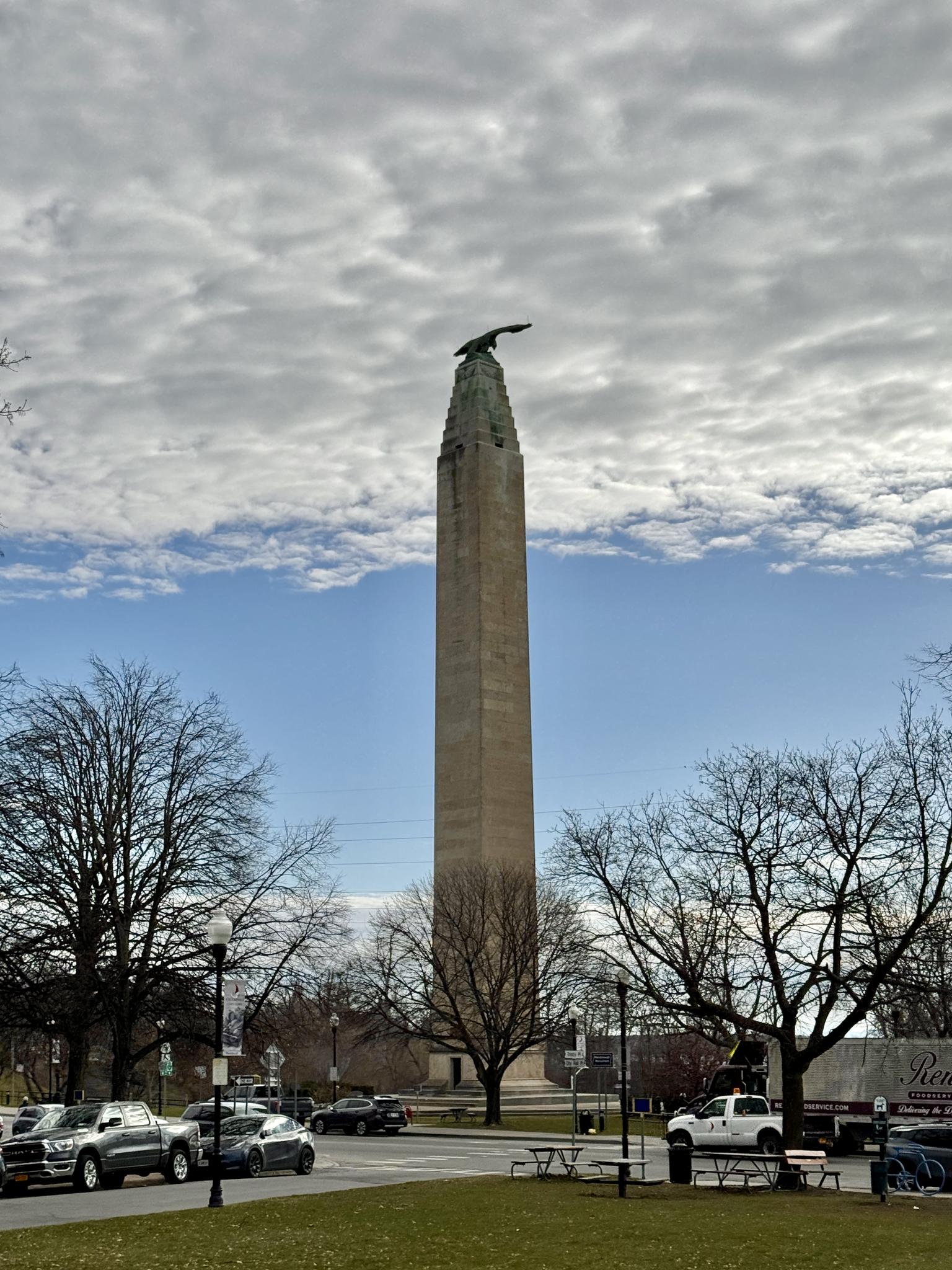 A tall obelisk monument with an eagle stands against a cloudy blue sky.