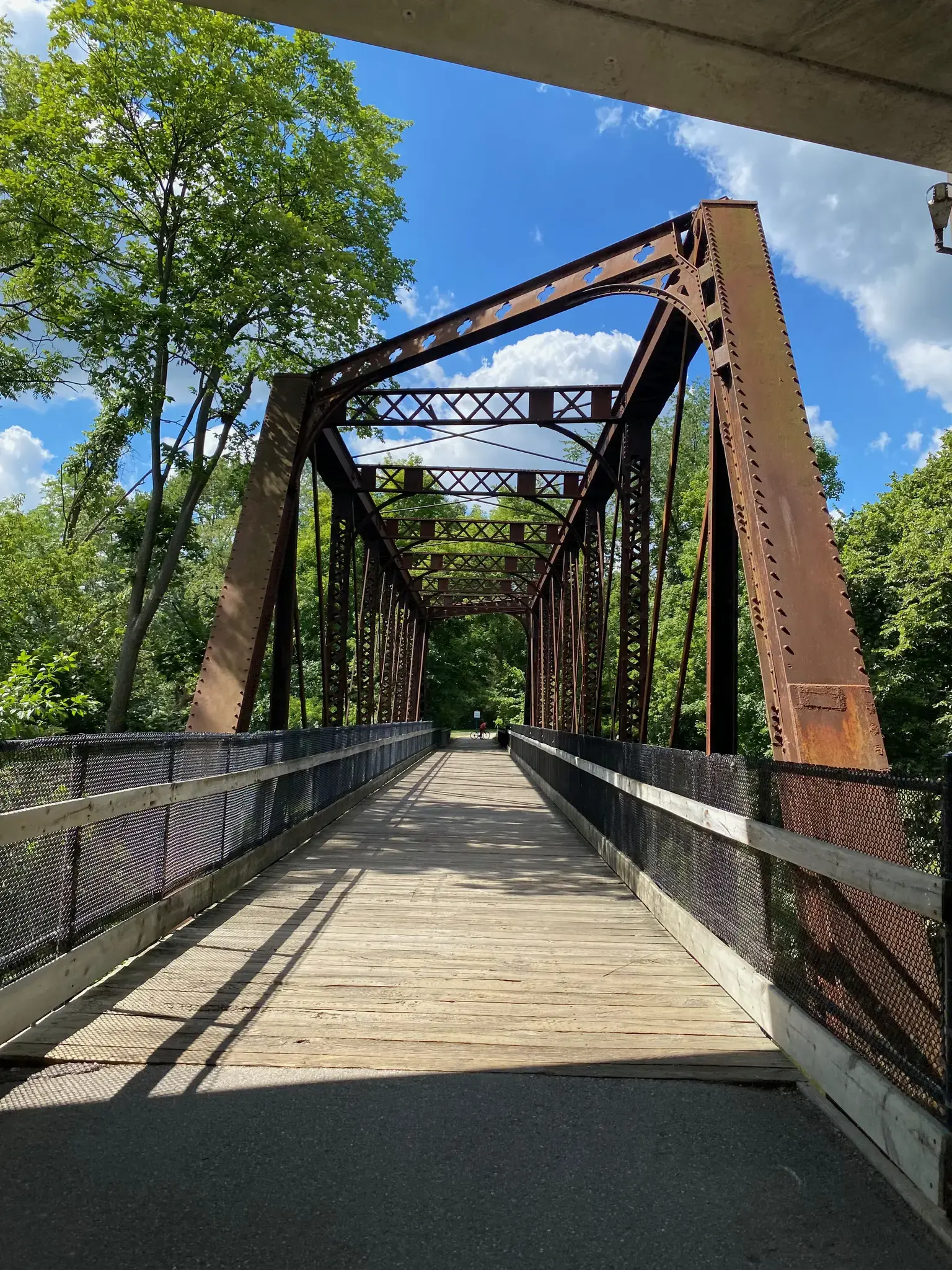 A long, rusty metal truss bridge with a wooden deck under a sunny sky.