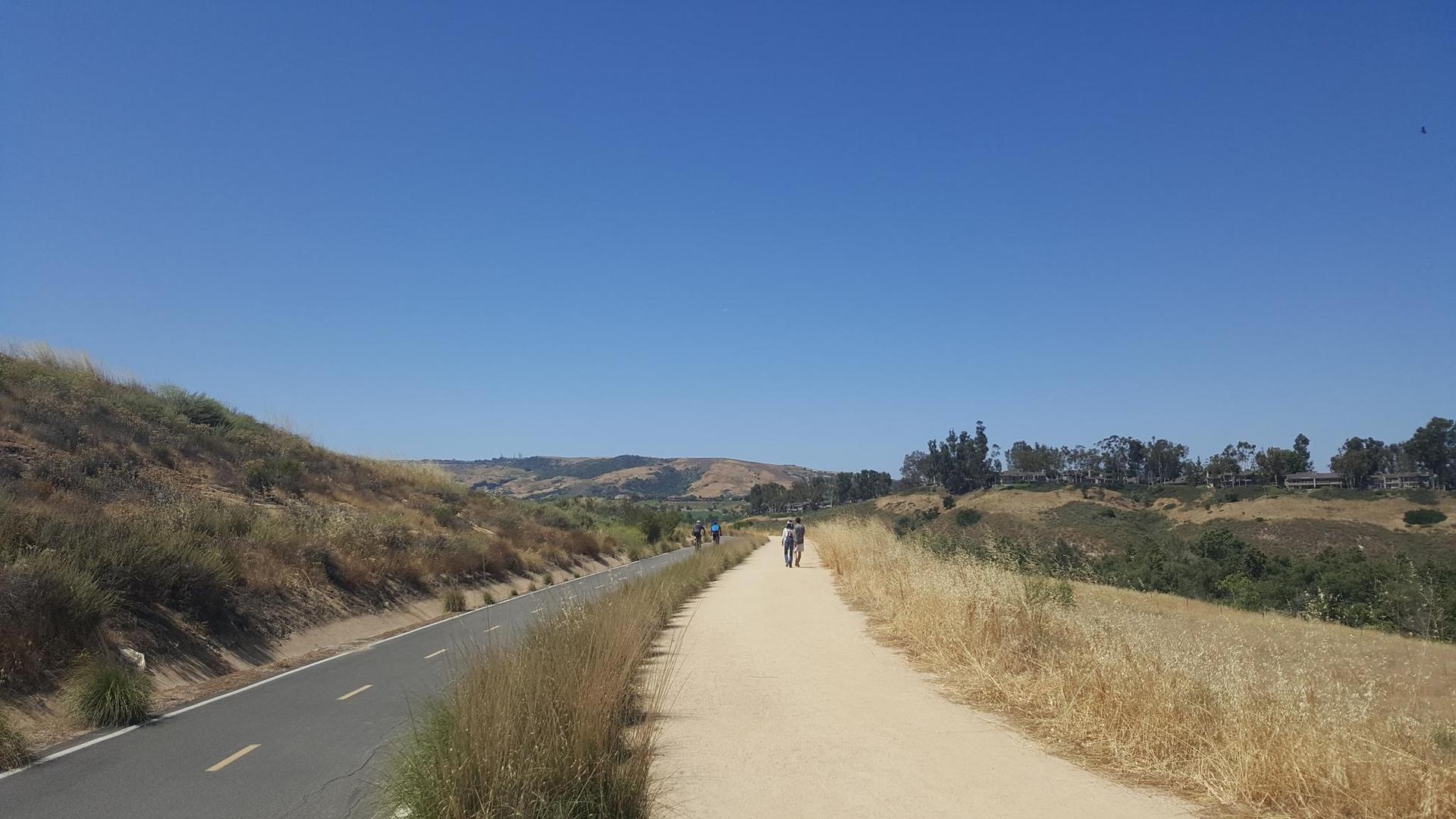 People walking on a dirt path next to a paved road through dry hills.