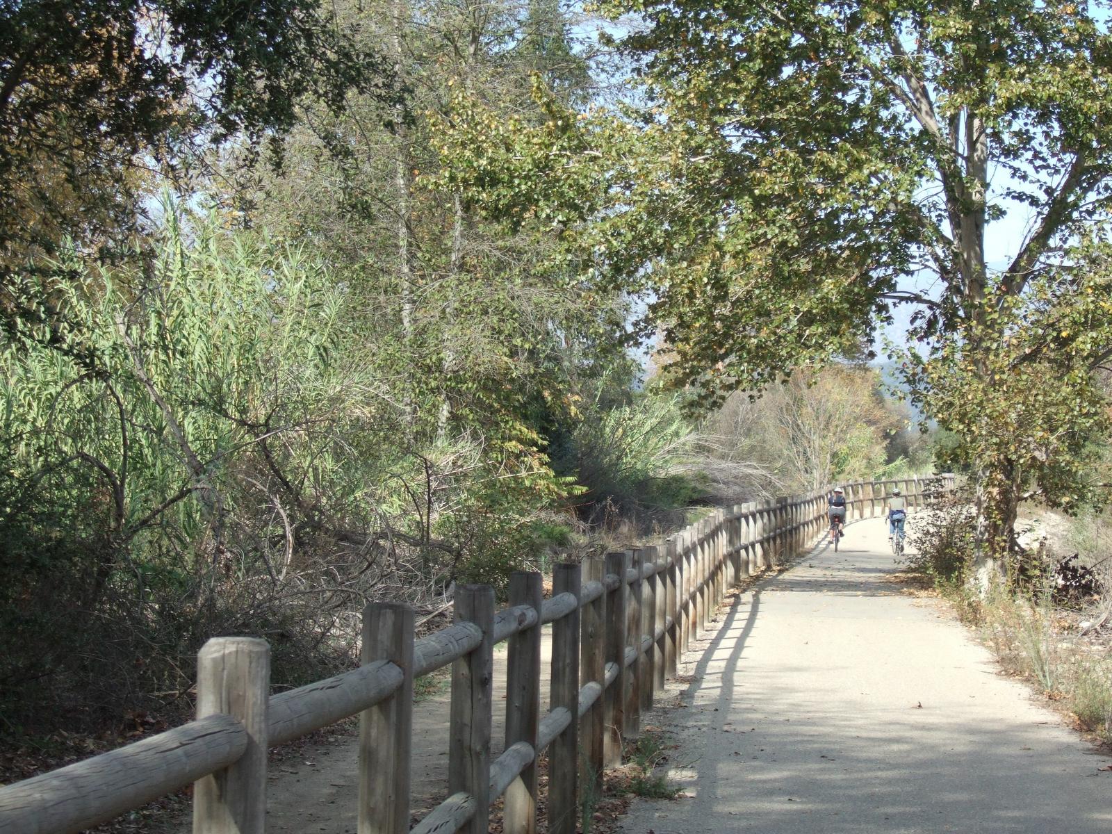 Sunny dirt path with a wooden fence, lush trees, and two distant cyclists.