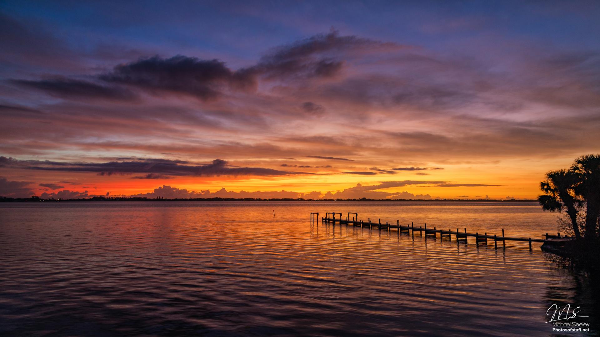 A long wooden pier extends into calm water under a vibrant, colorful sunset sky.