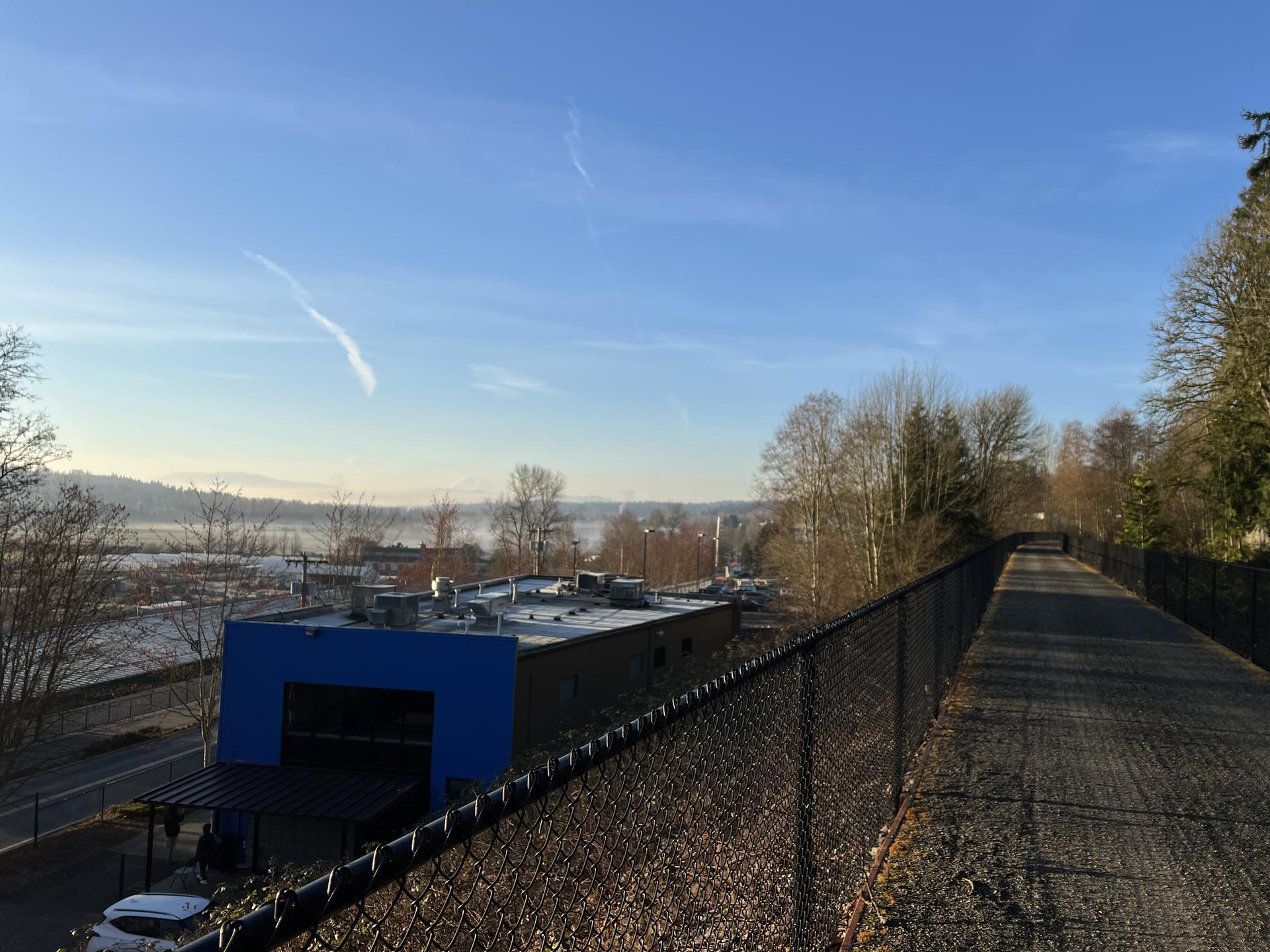 Elevated paved path next to a fence, overlooking industrial buildings and misty landscape.