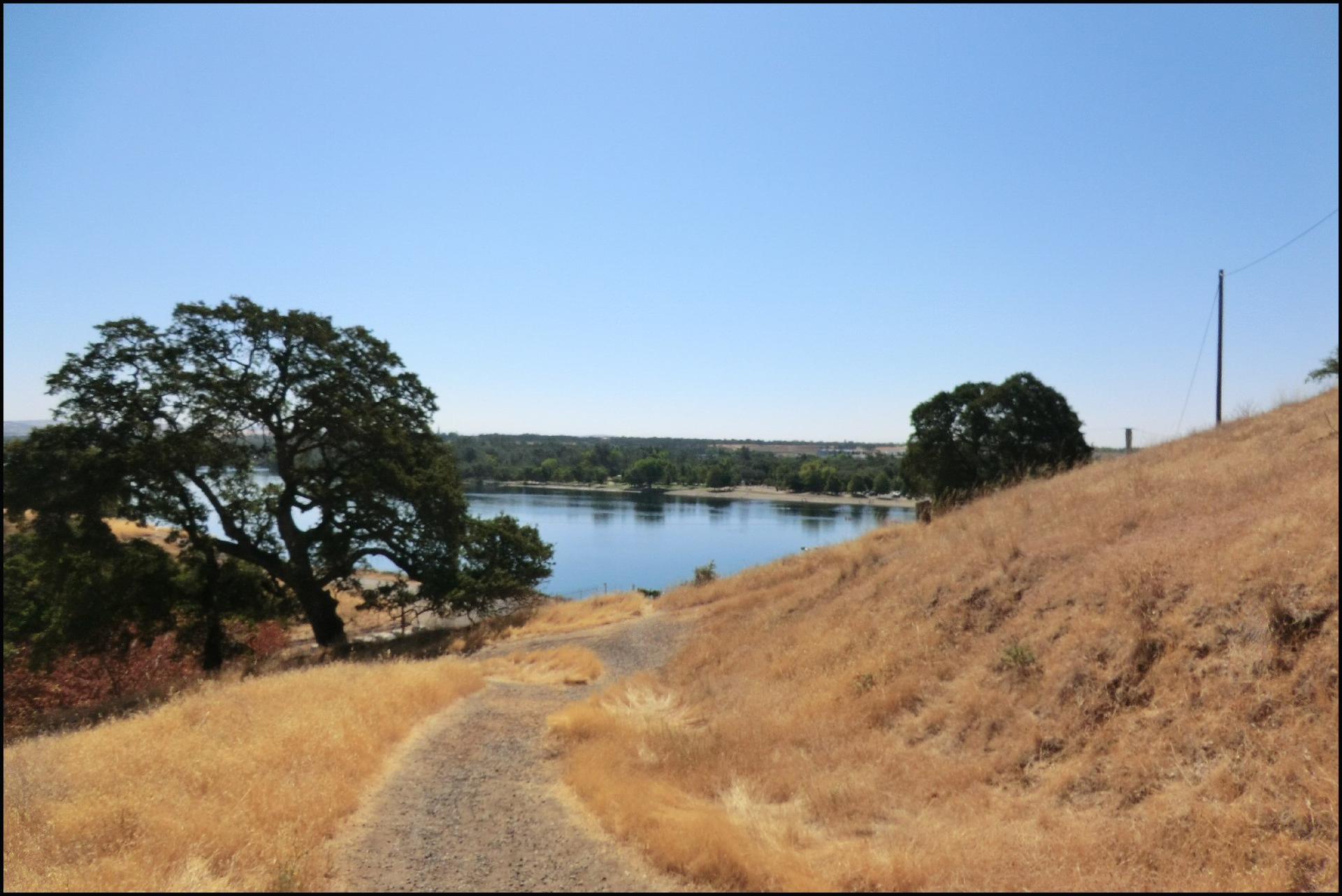 A winding dirt path descends a golden hillside towards a calm blue lake.