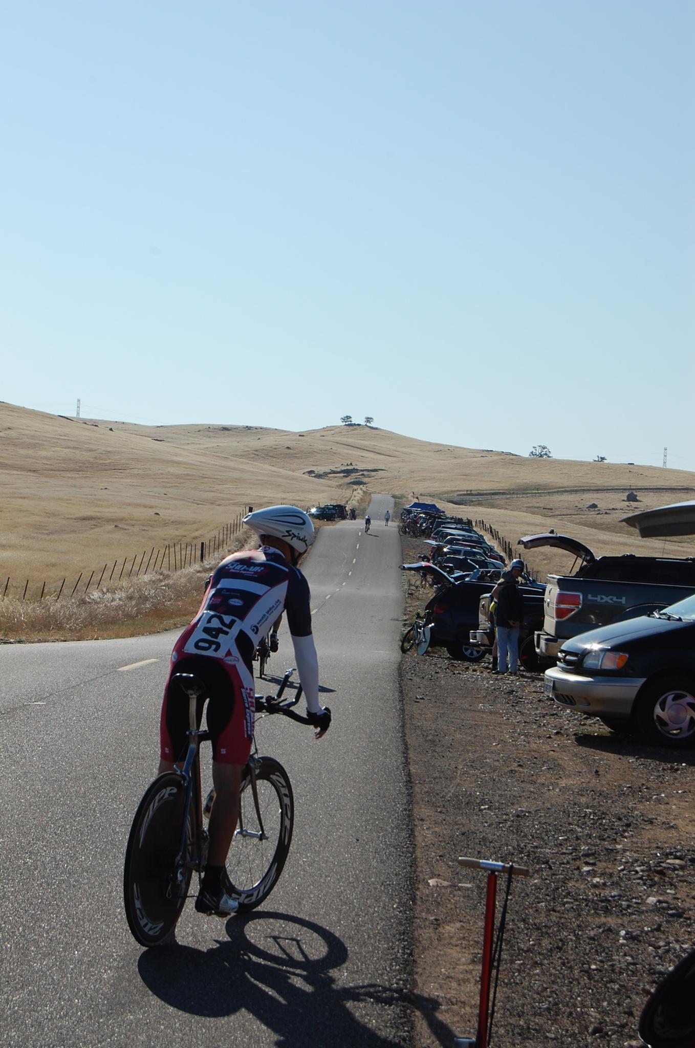 A cyclist rides uphill on a sunny road past parked cars.