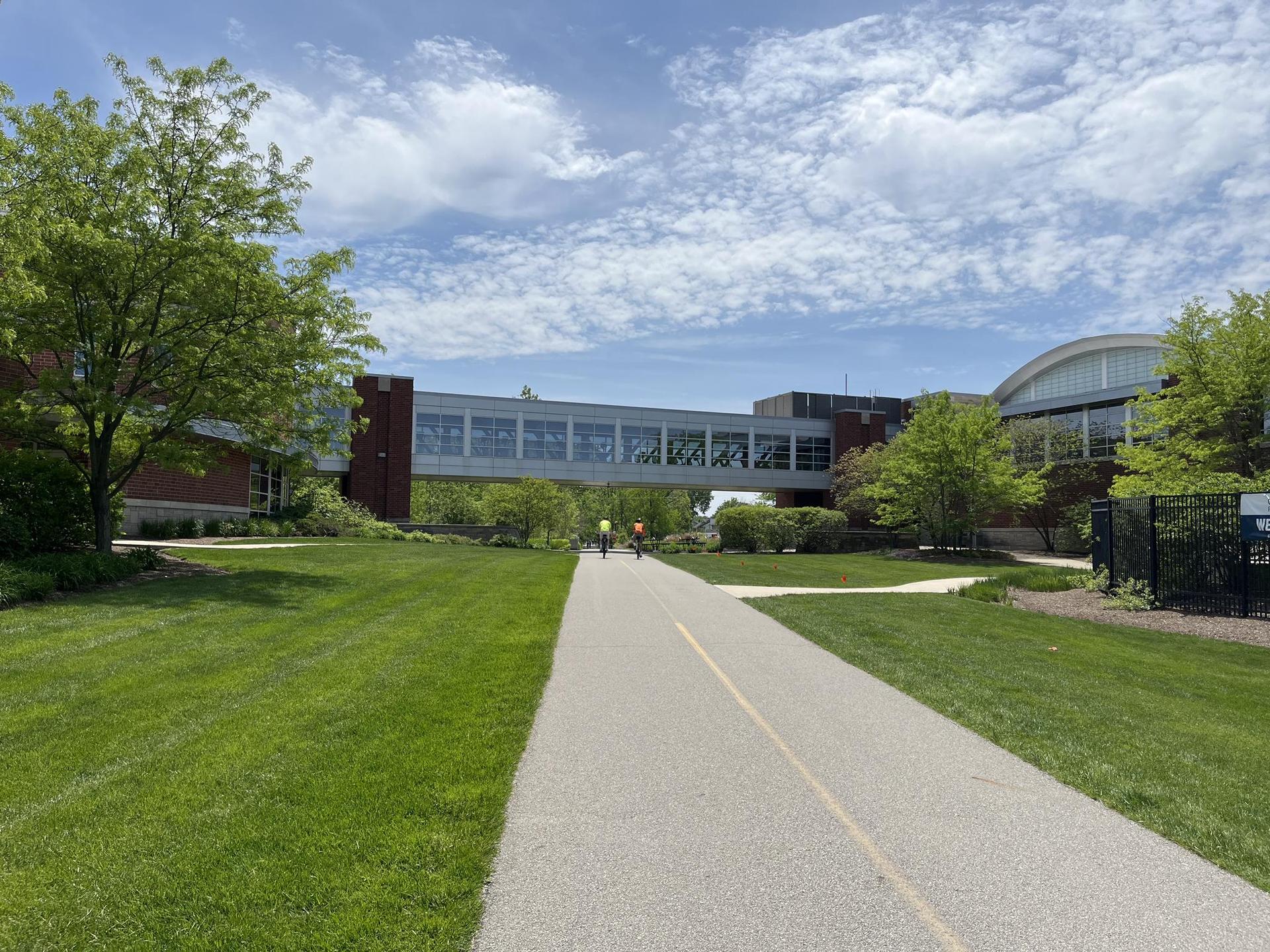 Paved path leading to a modern building with a skybridge, people biking.
